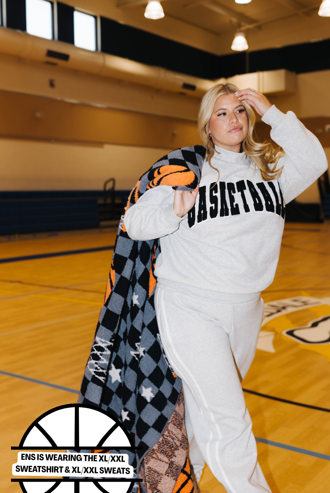 A woman posing in a gym on a basketball court wearing a gray sweatshirt and sweatpants set, with a black, gray, and orange checkered blanket draped over her shoulder. The sweatshirt features the word 'BASKETBALL' printed across the chest in black.