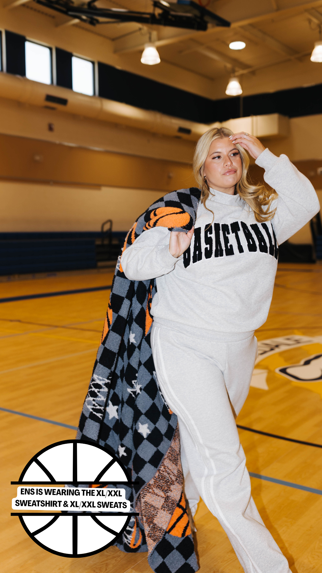 A woman posing in a gym on a basketball court wearing a gray sweatshirt and sweatpants set, with a black, gray, and orange checkered blanket draped over her shoulder. The sweatshirt features the word 'BASKETBALL' printed across the chest in black.
