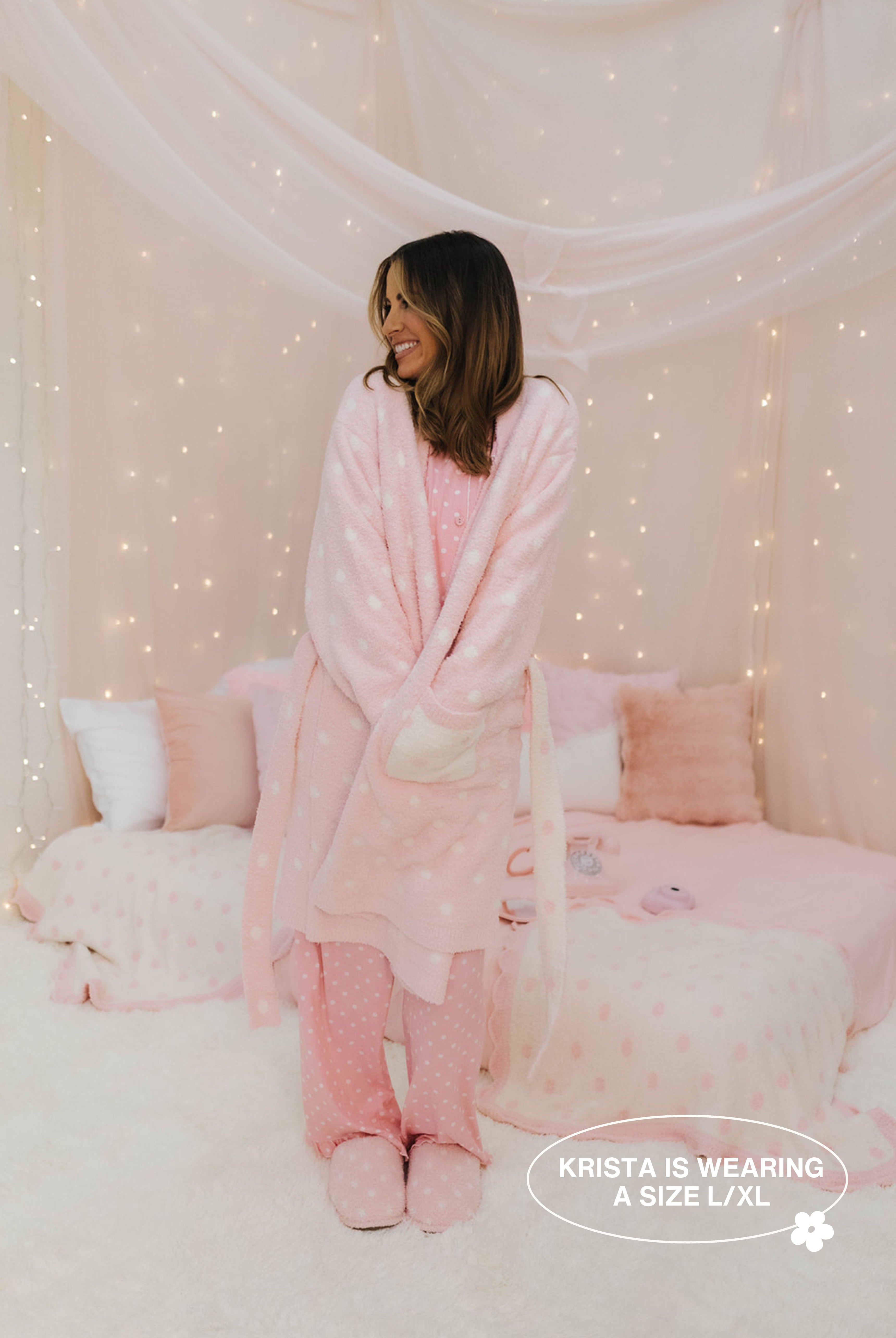 Woman wearing a pink plush robe and pajama set in a cozy room with fairy lights.