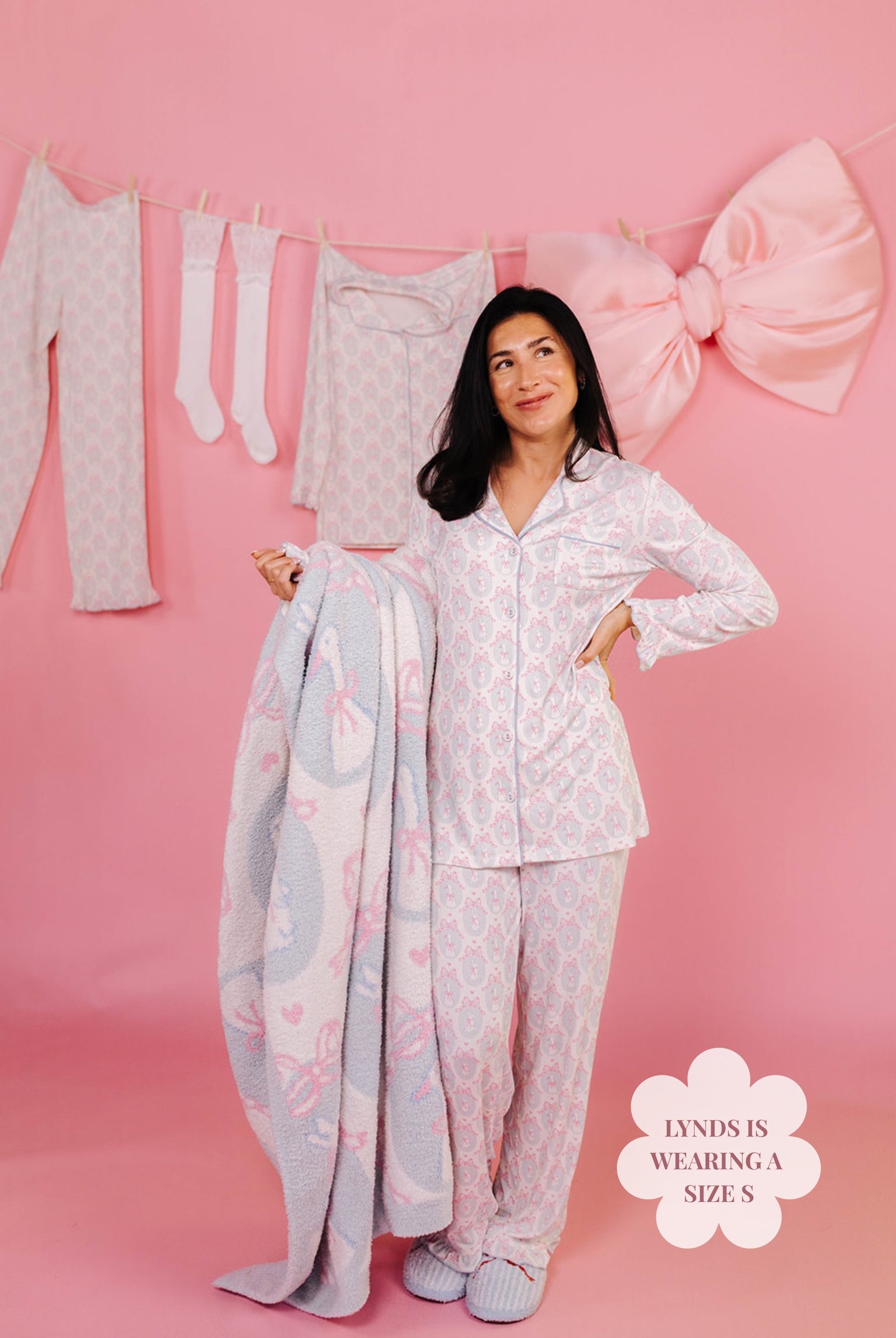 Woman in white pajama set holding a blanket with a large bow in the background.