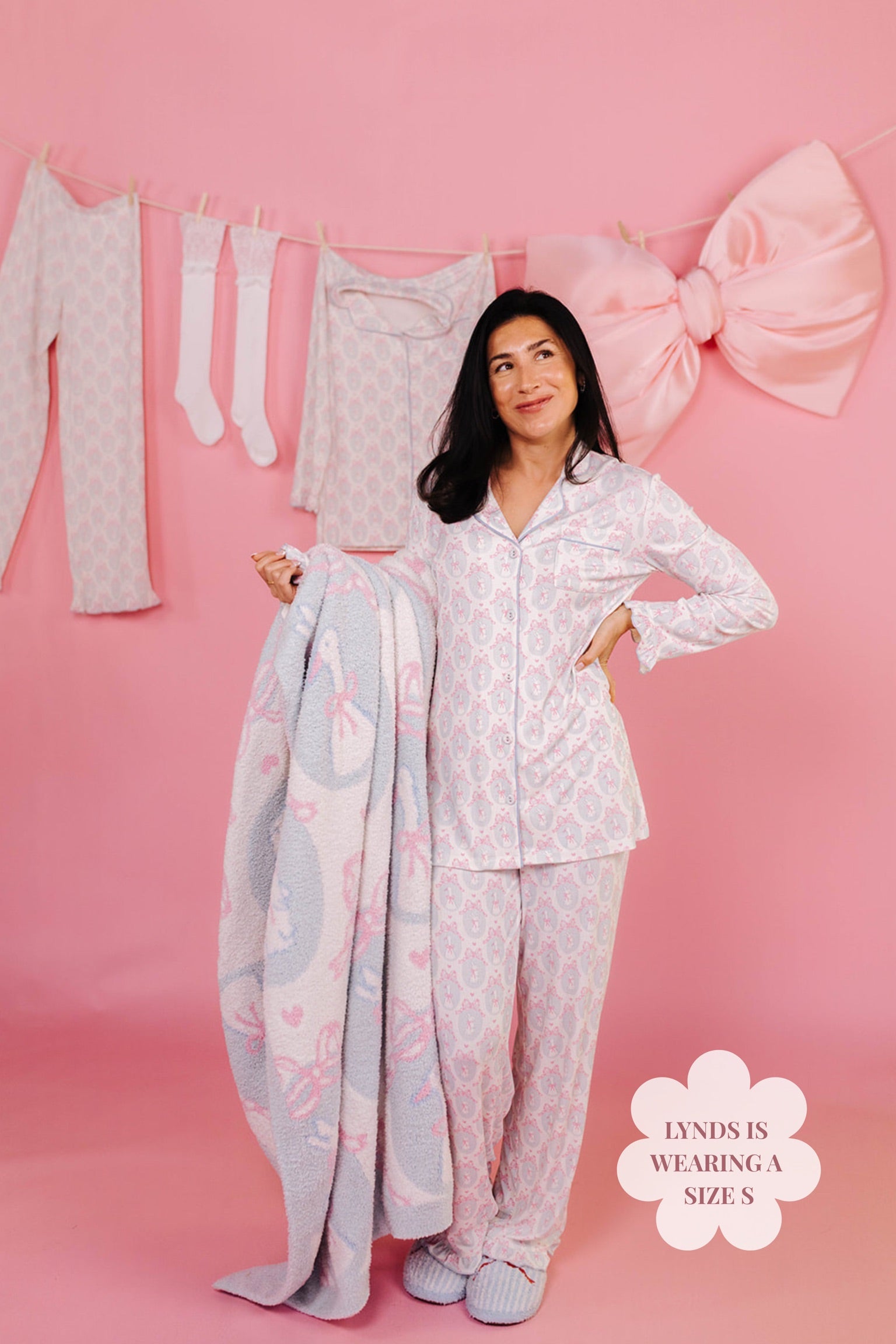 Woman in white pajama set holding a blanket with a large bow in the background.