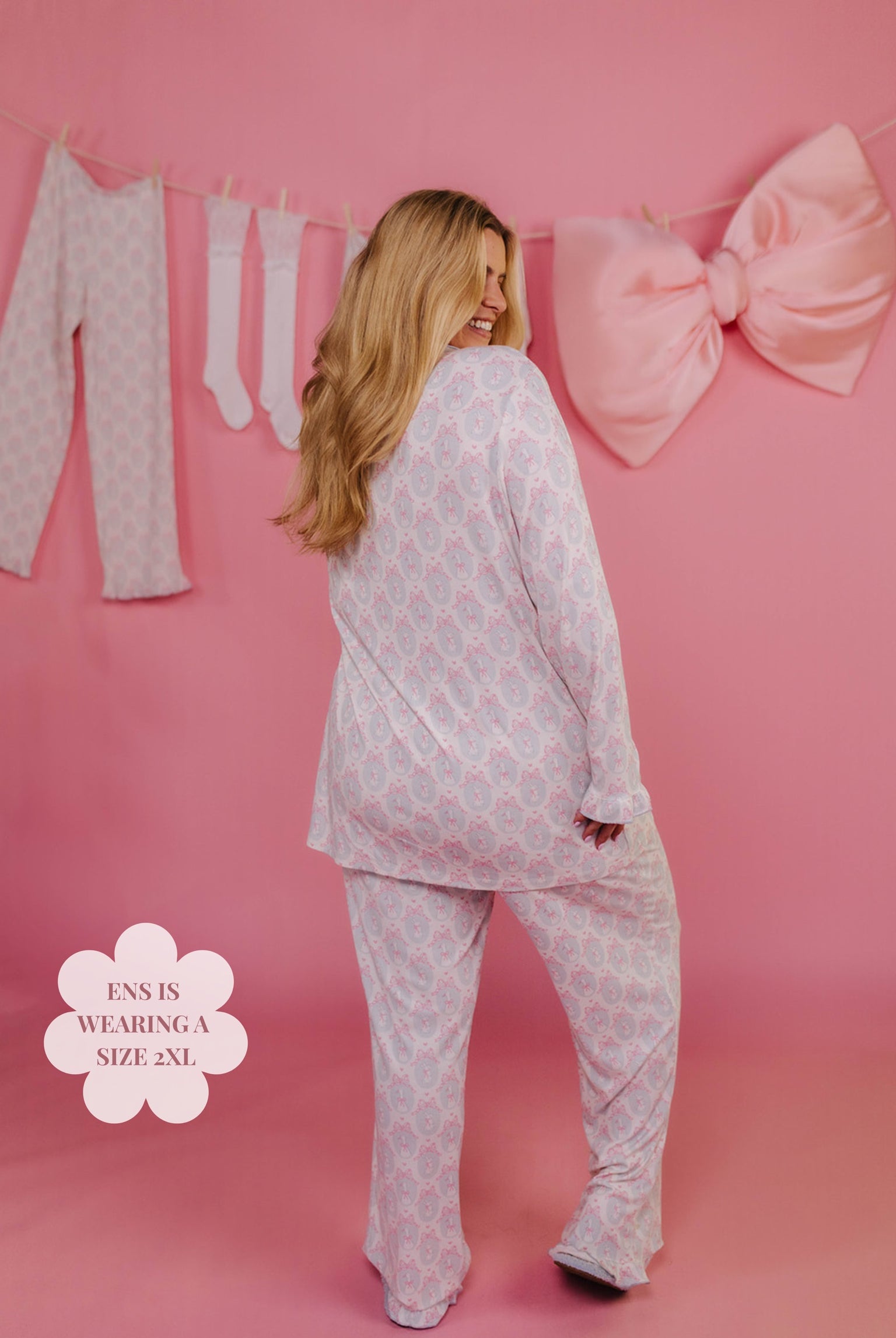 Woman wearing a white pajama set with a large bow and clothes in the background on a pink wall.
