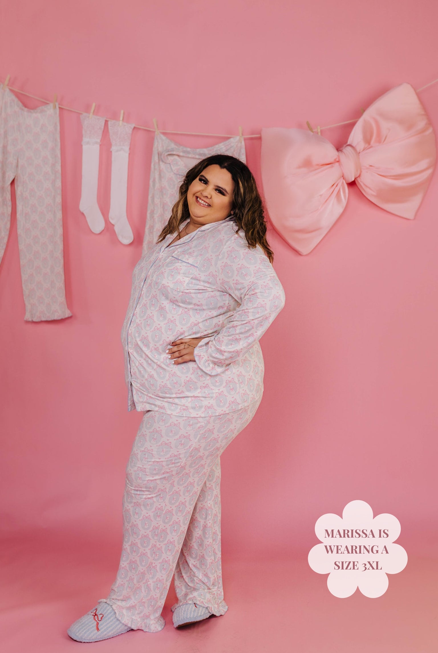 Woman wearing a white pajama set against a pink background with the pajamas hung and a big pink bow.