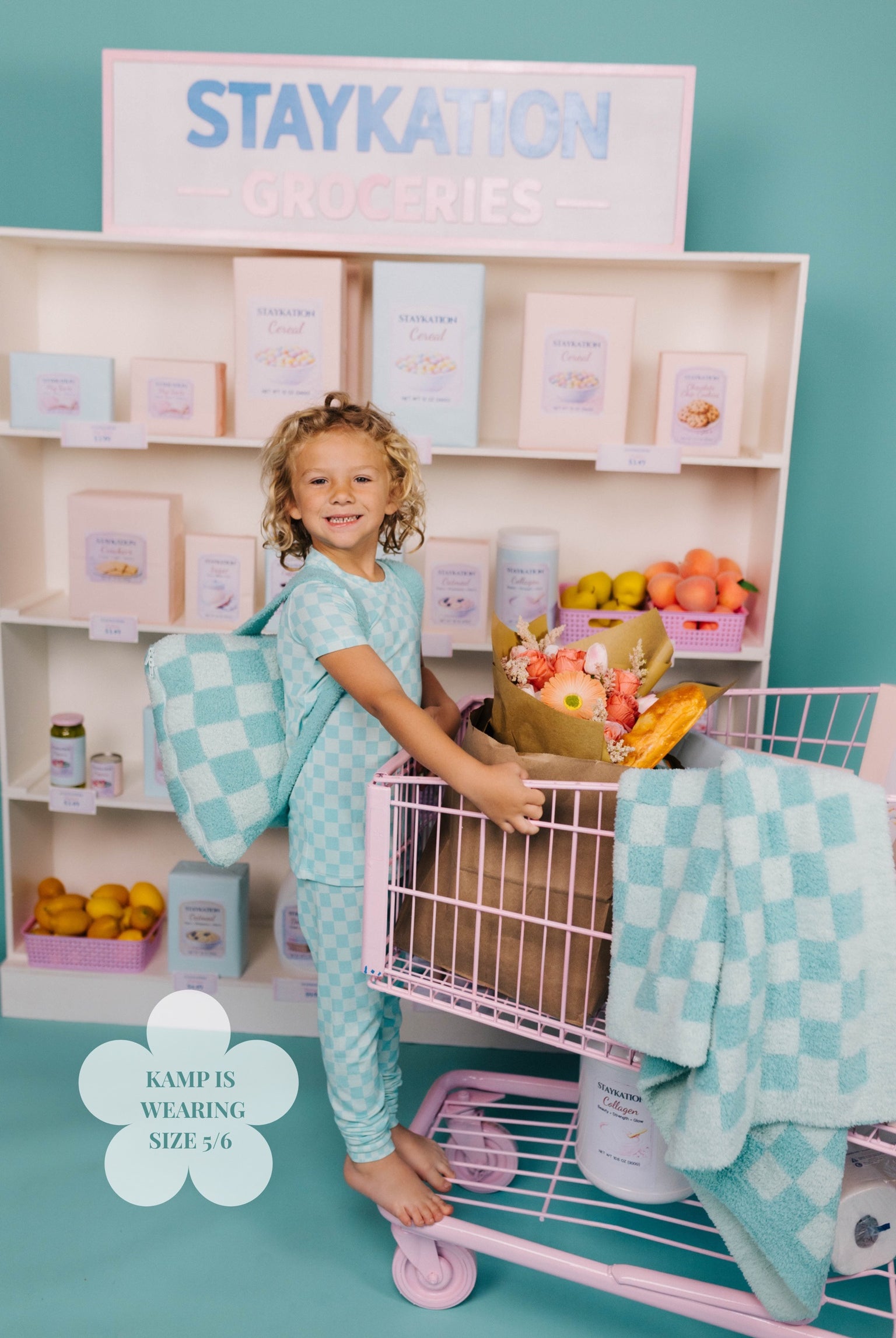 Child in teal pajamas with a checkered pattern standing next to a display of pretend Staykation products.