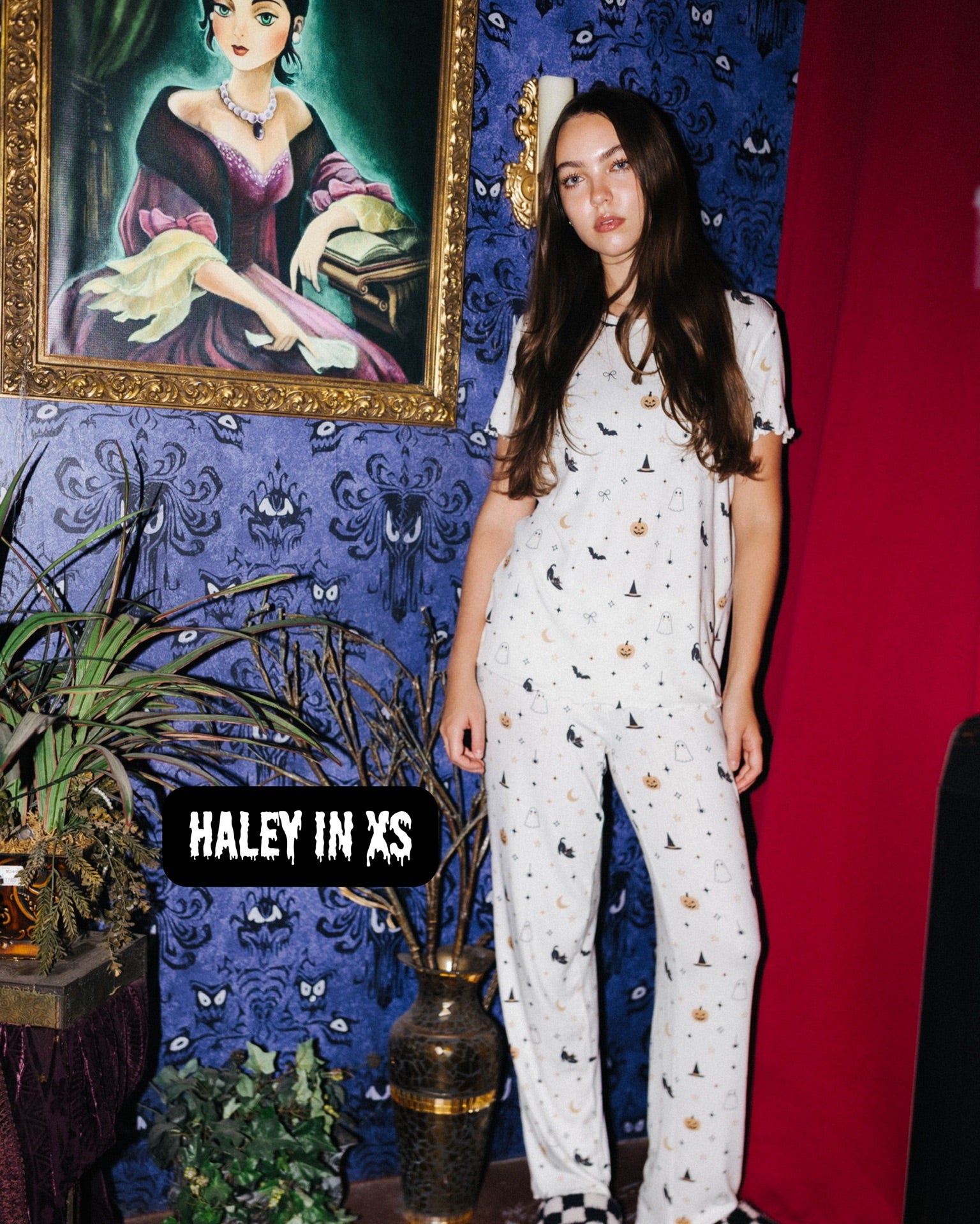 A woman stands in a styled room wearing a white short-sleeve pajama set with small black and gold motifs.