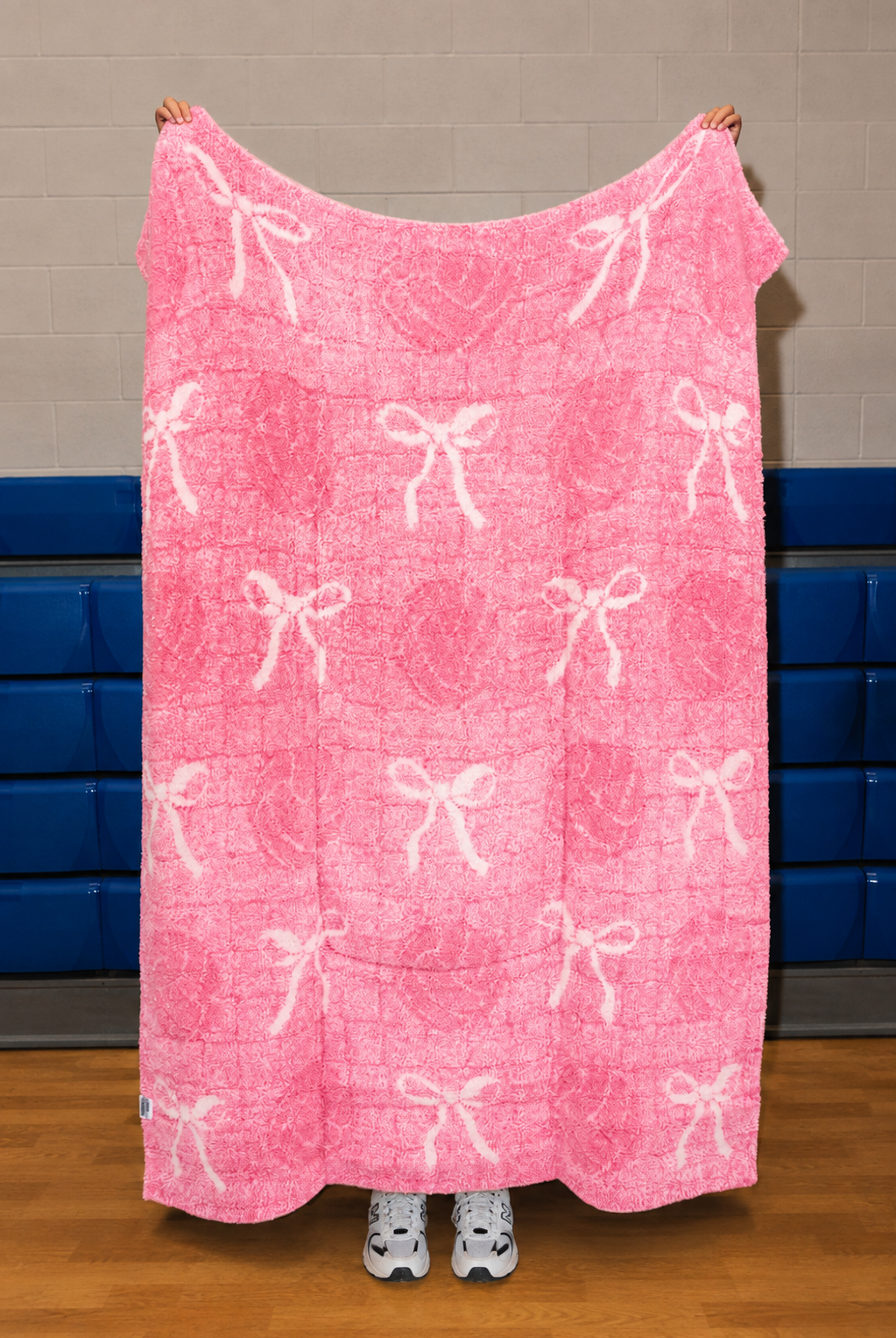 A pink blanket with a white bow and heart pattern is held up by a person in a gym with blue padding on the wall.