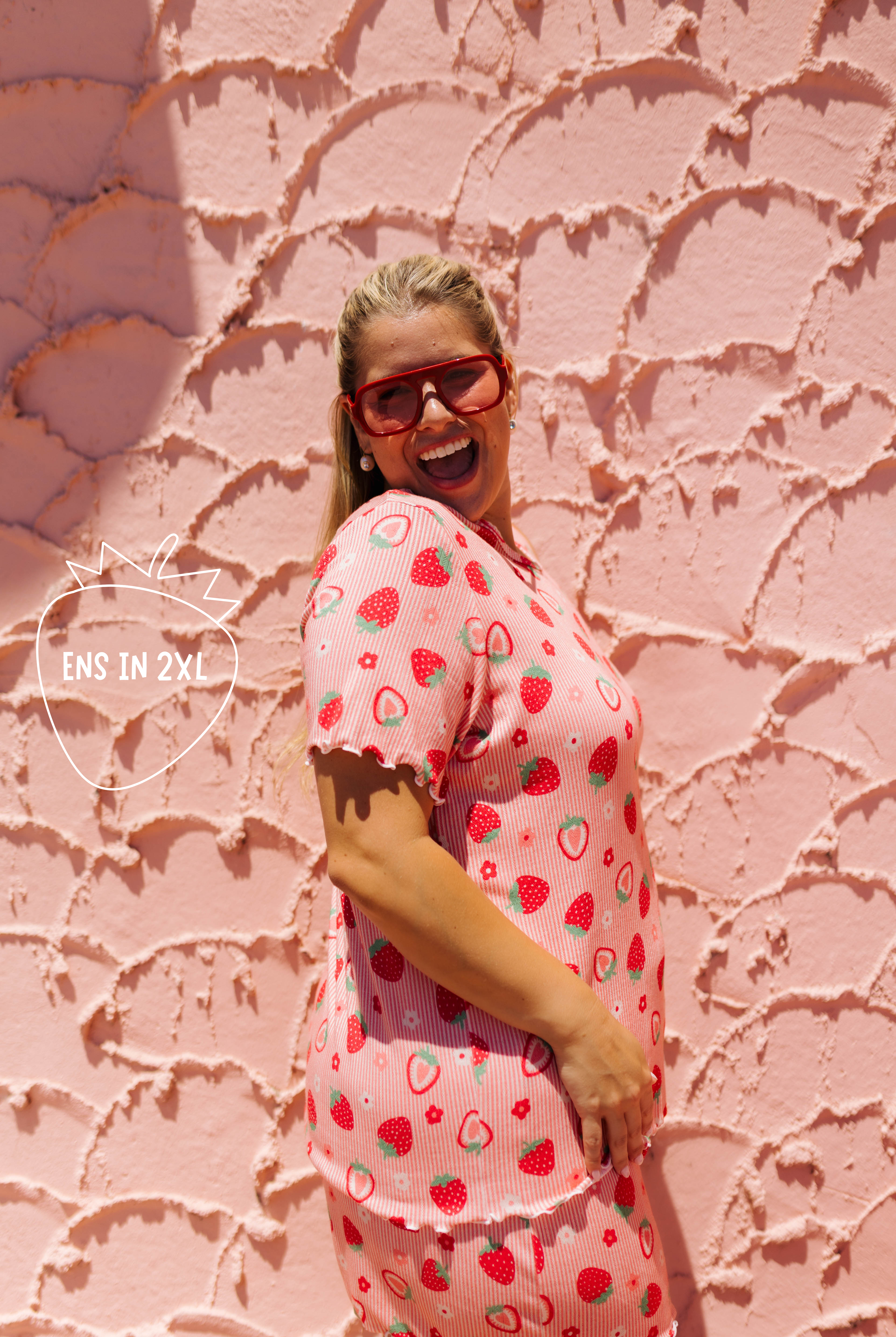 A woman wearing a pink strawberry-patterned short-sleeve top and matching shorts stands against a textured pink wall, smiling and wearing red-framed sunglasses. A watermark on the left reads 'ENS IN 2X1'.