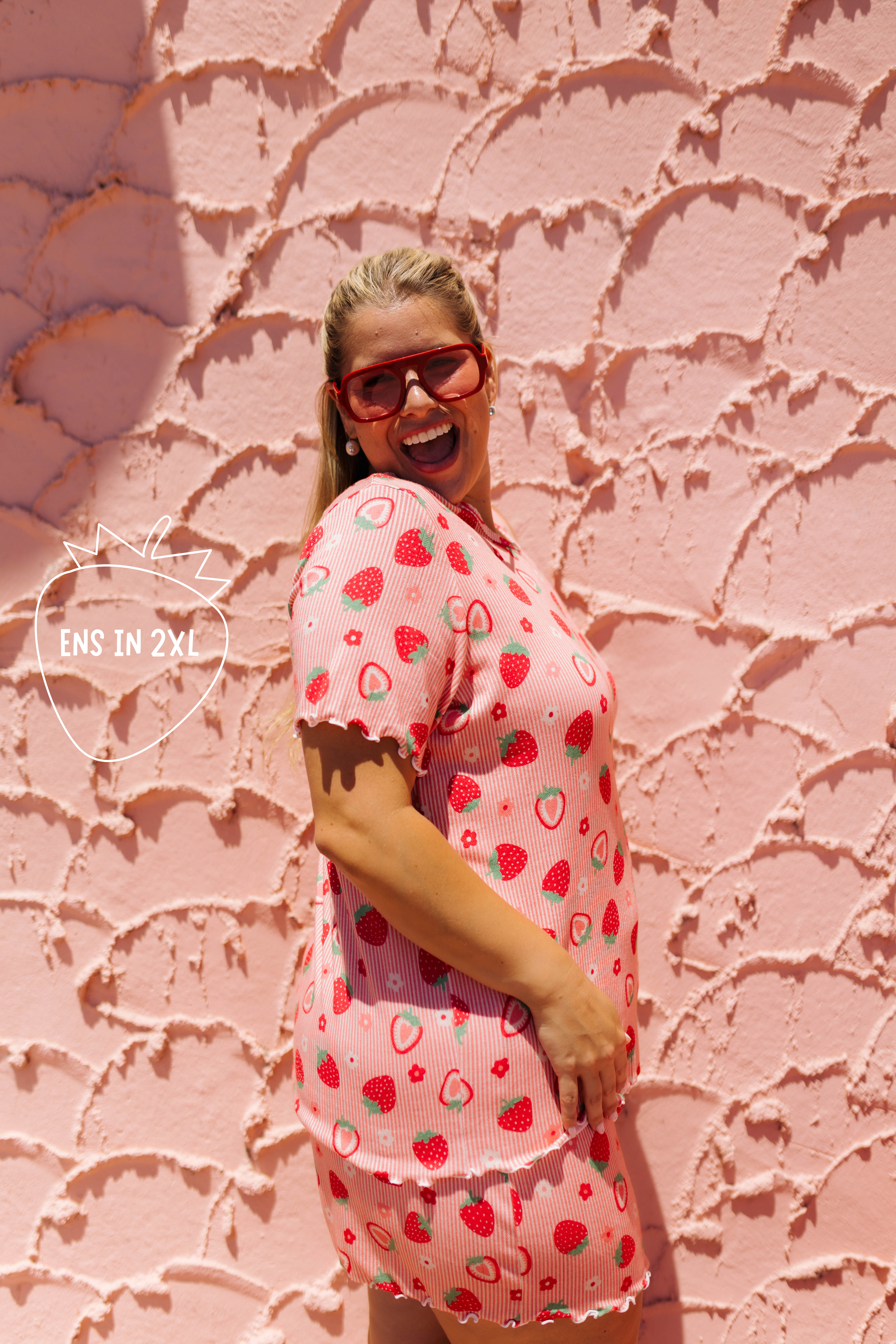 A woman wearing a pink strawberry-patterned short-sleeve top and matching shorts stands against a textured pink wall, smiling and wearing red-framed sunglasses. A watermark on the left reads 'ENS IN 2X1'.