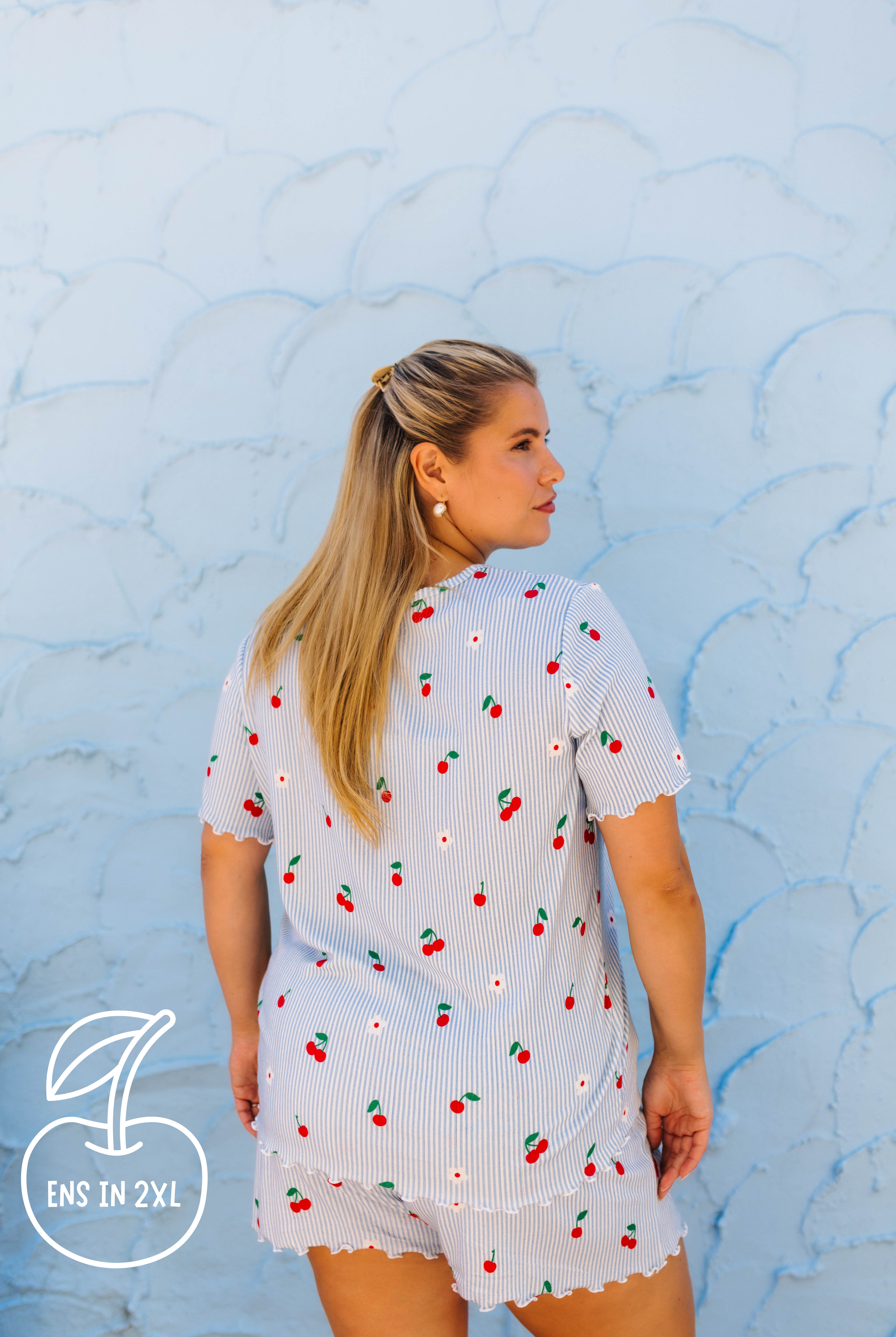 A woman wearing a light blue and white strawberry-print short-sleeve top and matching shorts, photographed from the back against a textured light blue wall.
