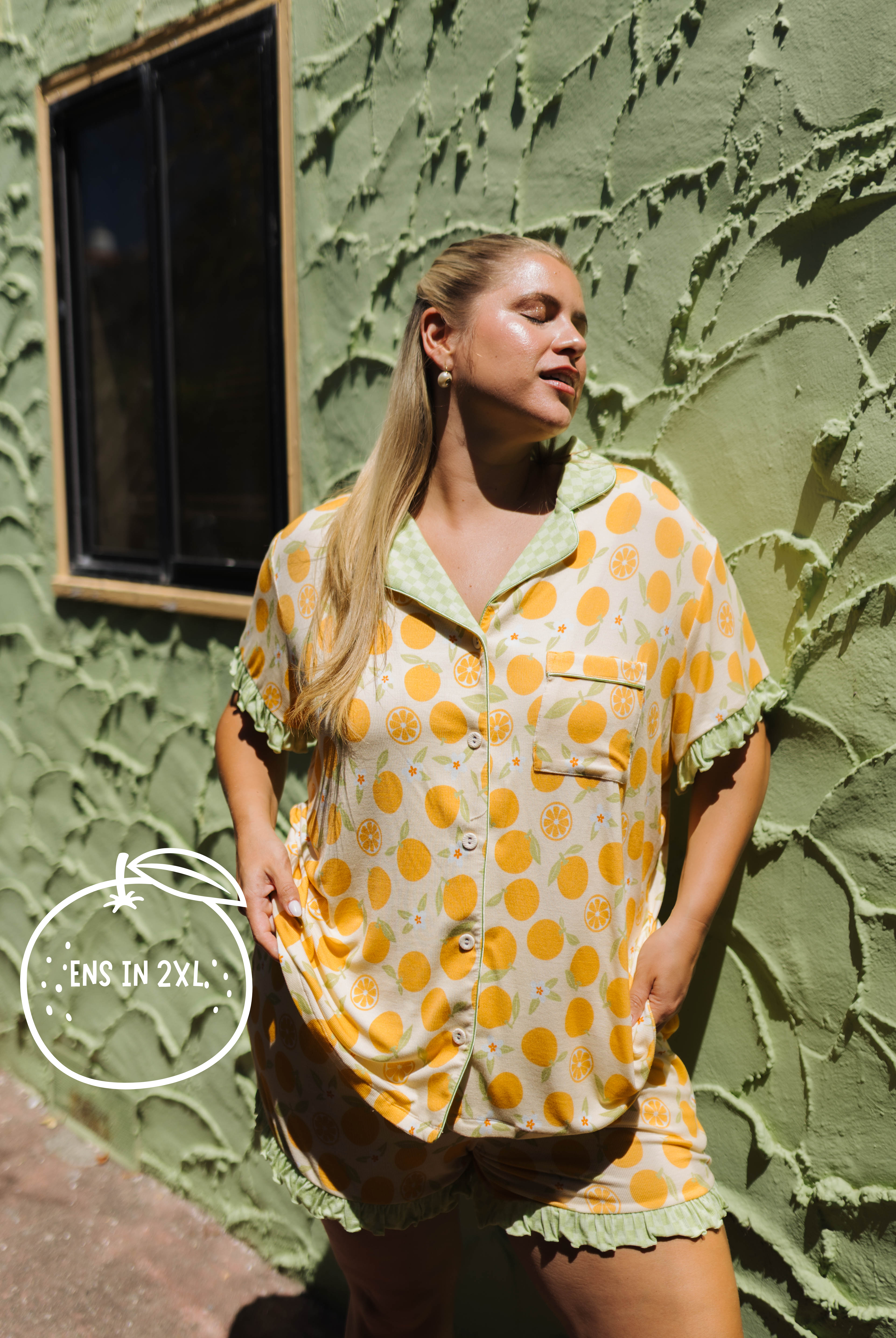 A woman modeling an orange citrus-patterned short-sleeve pajama set with green ruffled trim, standing against a textured olive green stucco wall.