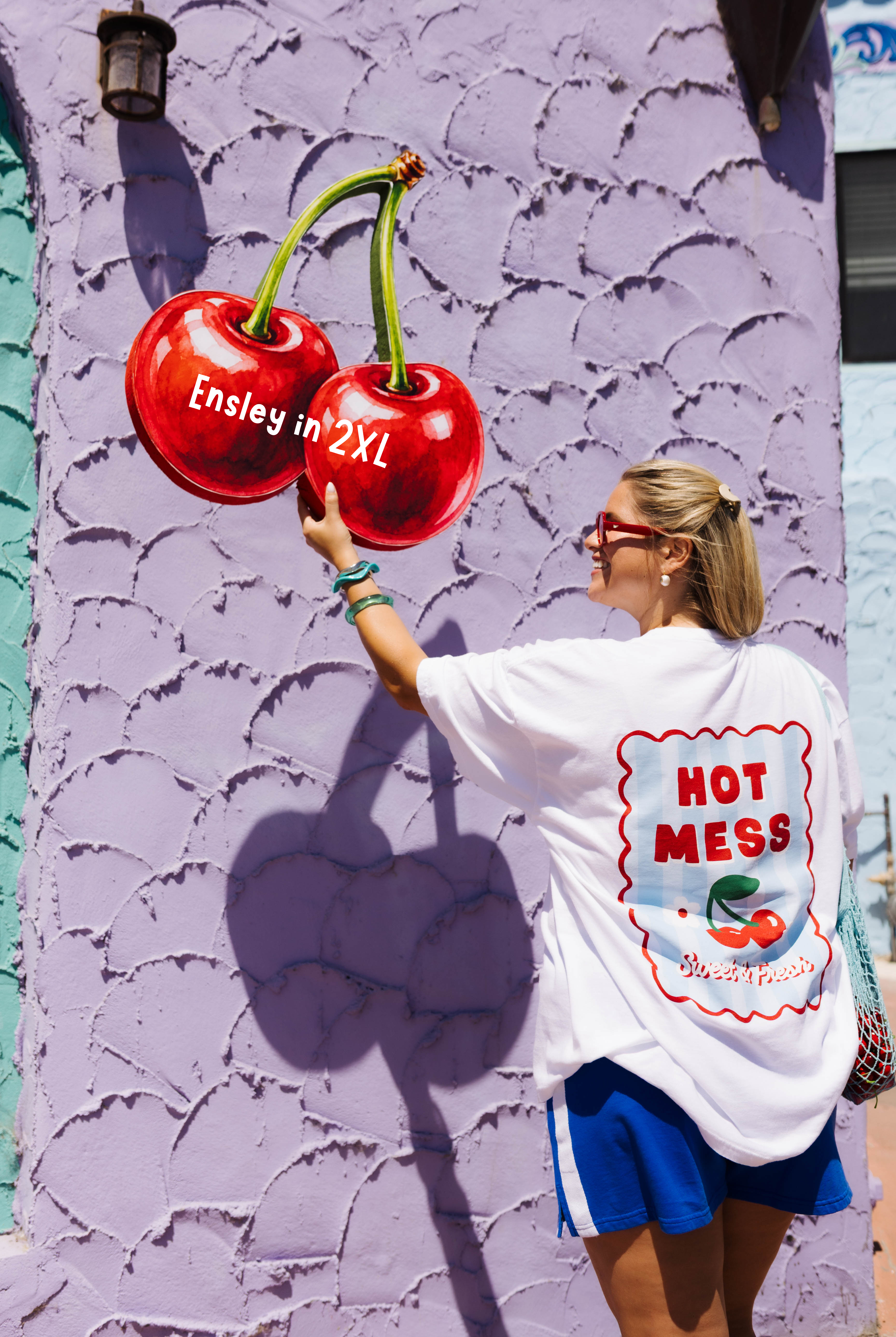 A woman wearing a white graphic t-shirt and blue shorts stands in front of a textured lavender wall, reaching toward a large 3D cherry mural painted on the wall. The t-shirt features a large graphic on the back with the text 'HOT MESS' and 'Ensley in 2XL'.