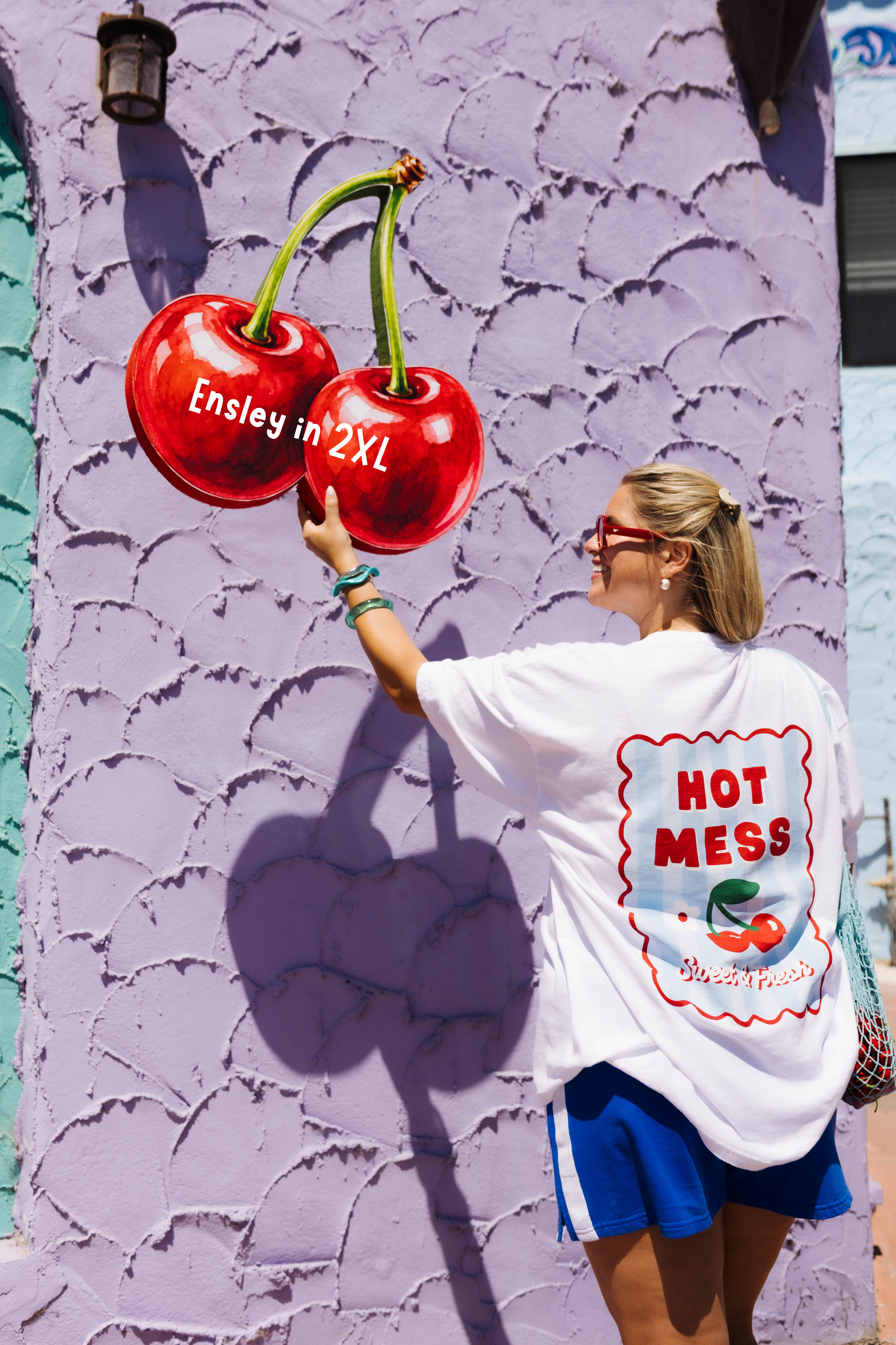 A woman wearing a white graphic t-shirt and blue shorts stands in front of a textured lavender wall, reaching toward a large 3D cherry mural painted on the wall. The t-shirt features a large graphic on the back with the text 'HOT MESS' and 'Ensley in 2XL'.