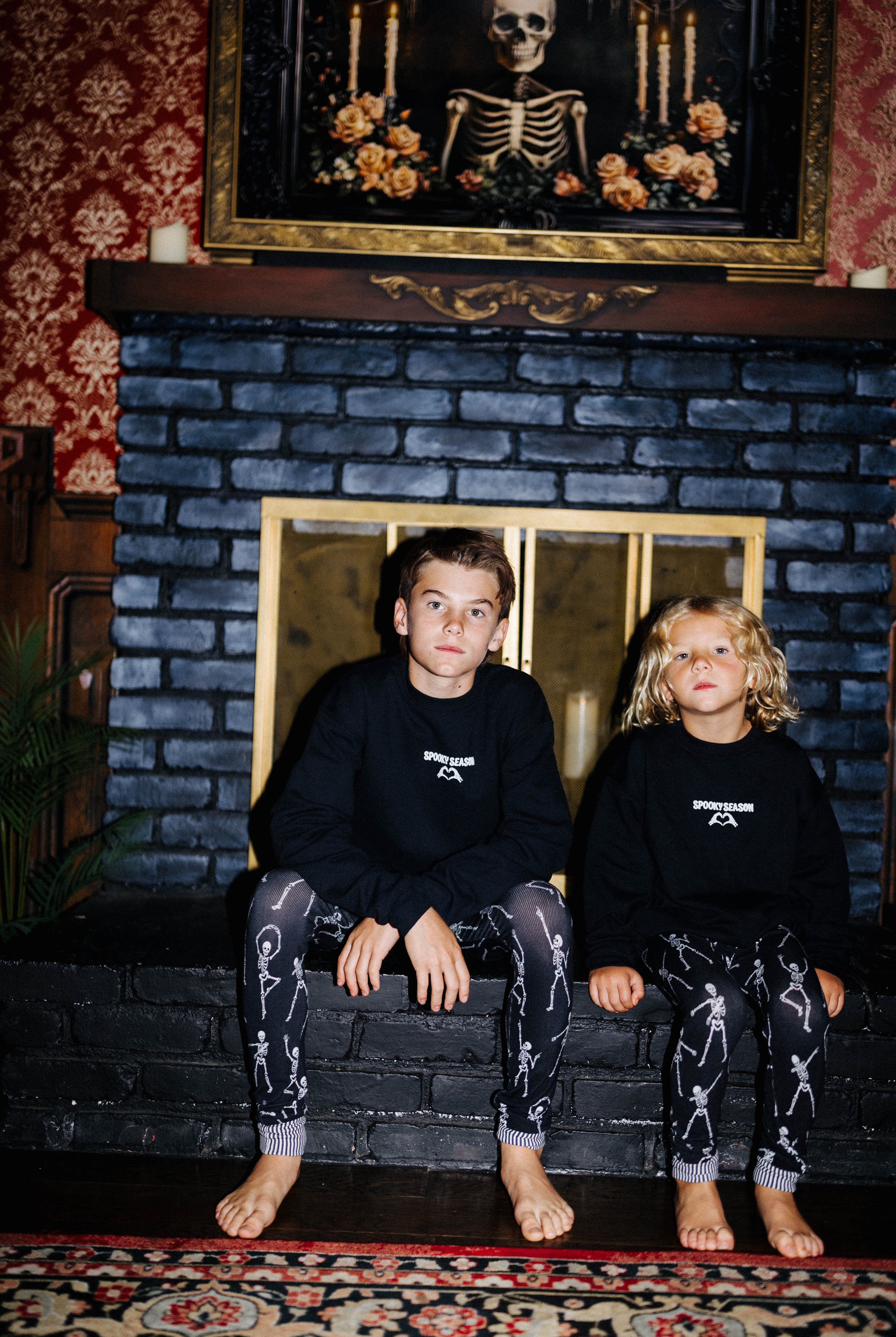 Two children wearing black Spooky Season sweatshirts by a fireplace