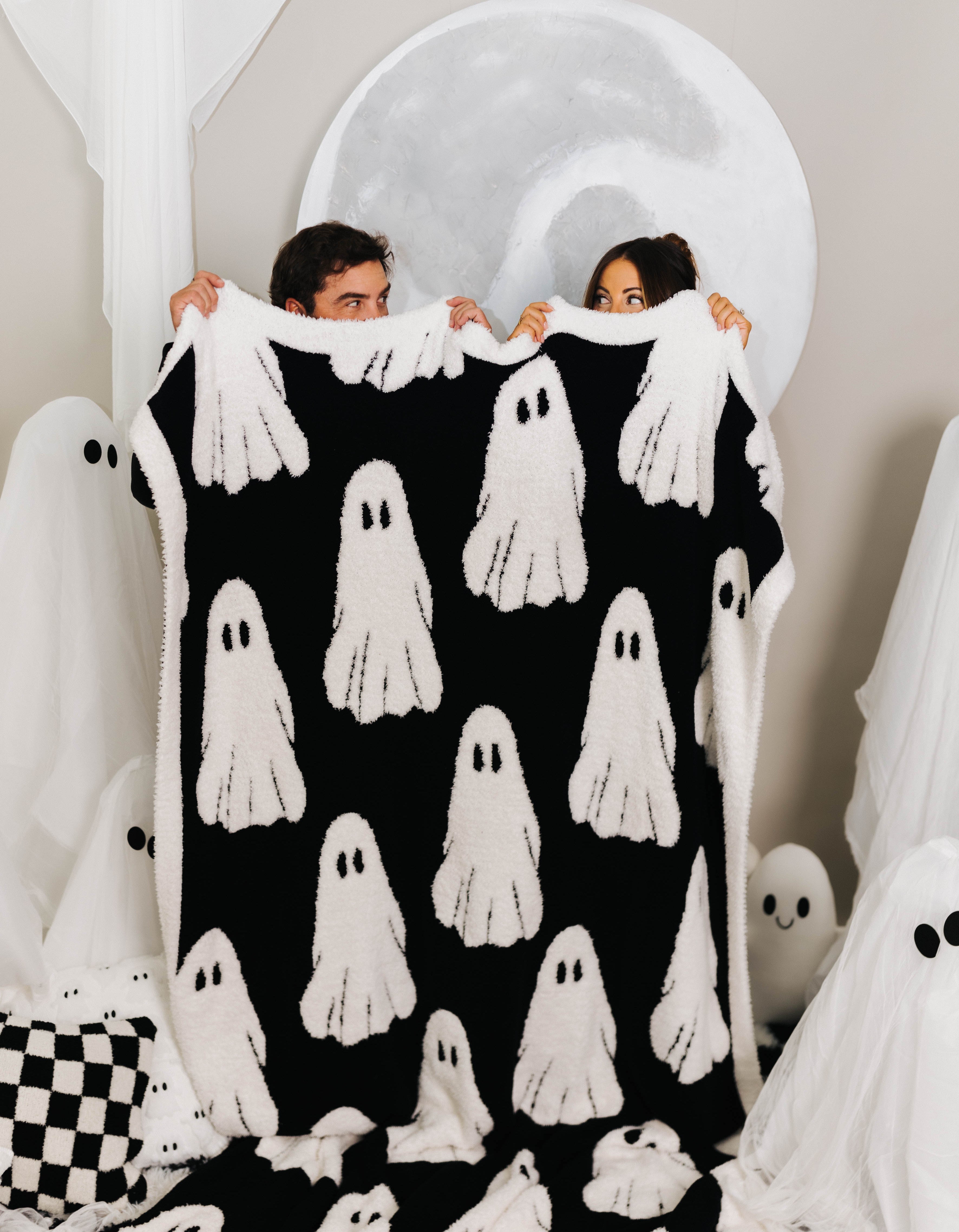 Two people holding up black and white ghost blanket looking at one another with ghost decorations behind them