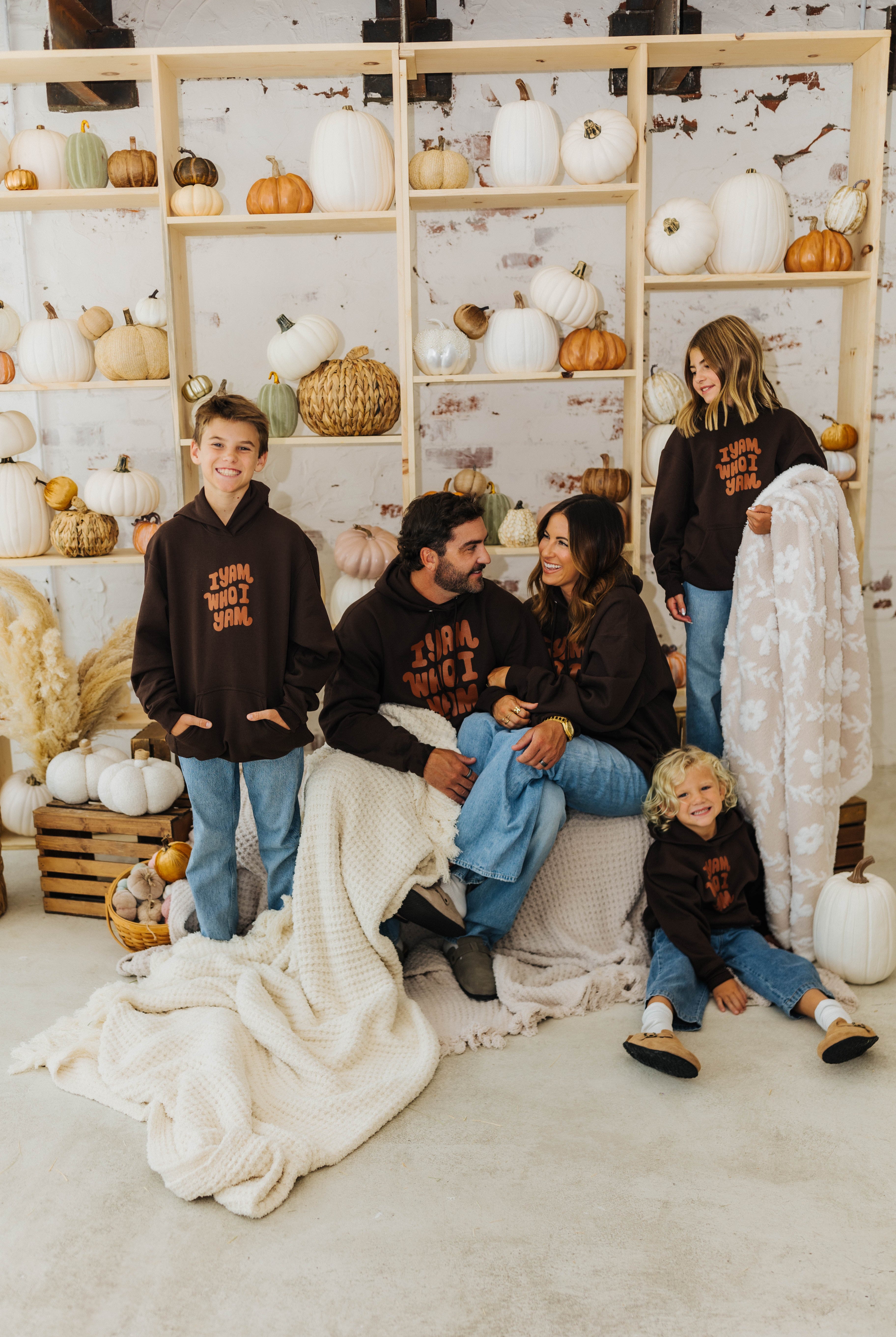 family smiling at each other sitting and standing on hay with waffle blankets and holding pumpkin blanket with pumpkin decorations in the background