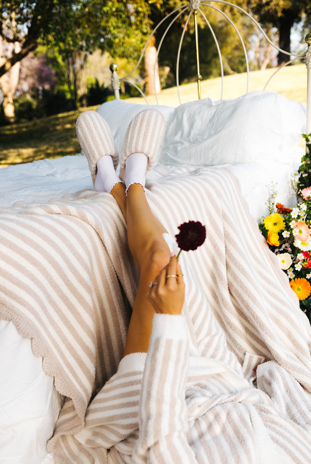 Person lying on a white blanket outdoors with striped pants and white socks, holding a flower.