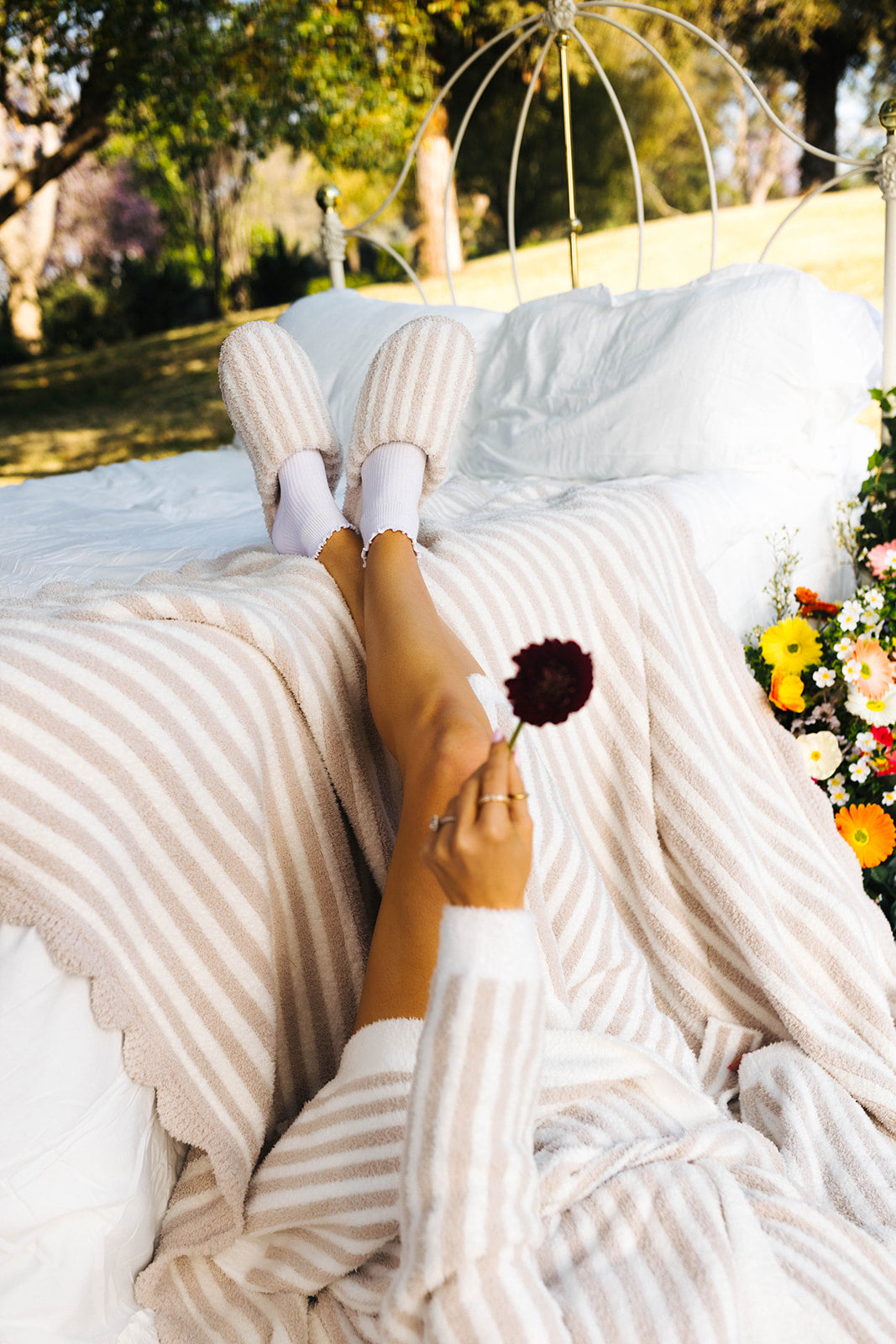 Person lying on a white blanket outdoors with striped pants and white socks, holding a flower.