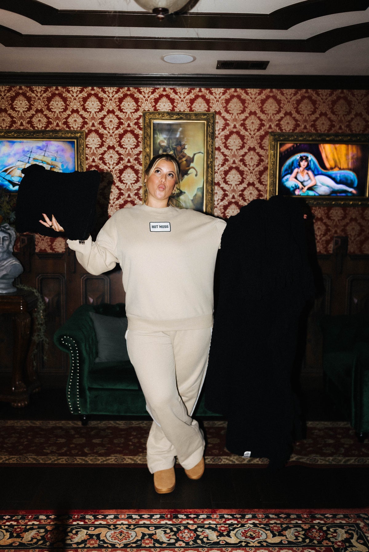 A woman stands in a vintage-style room with patterned wallpaper and framed paintings, holding up a black sweatshirt and a black jacket while wearing a cream sweatshirt and matching sweatpants. A small tag is visible on her chest.