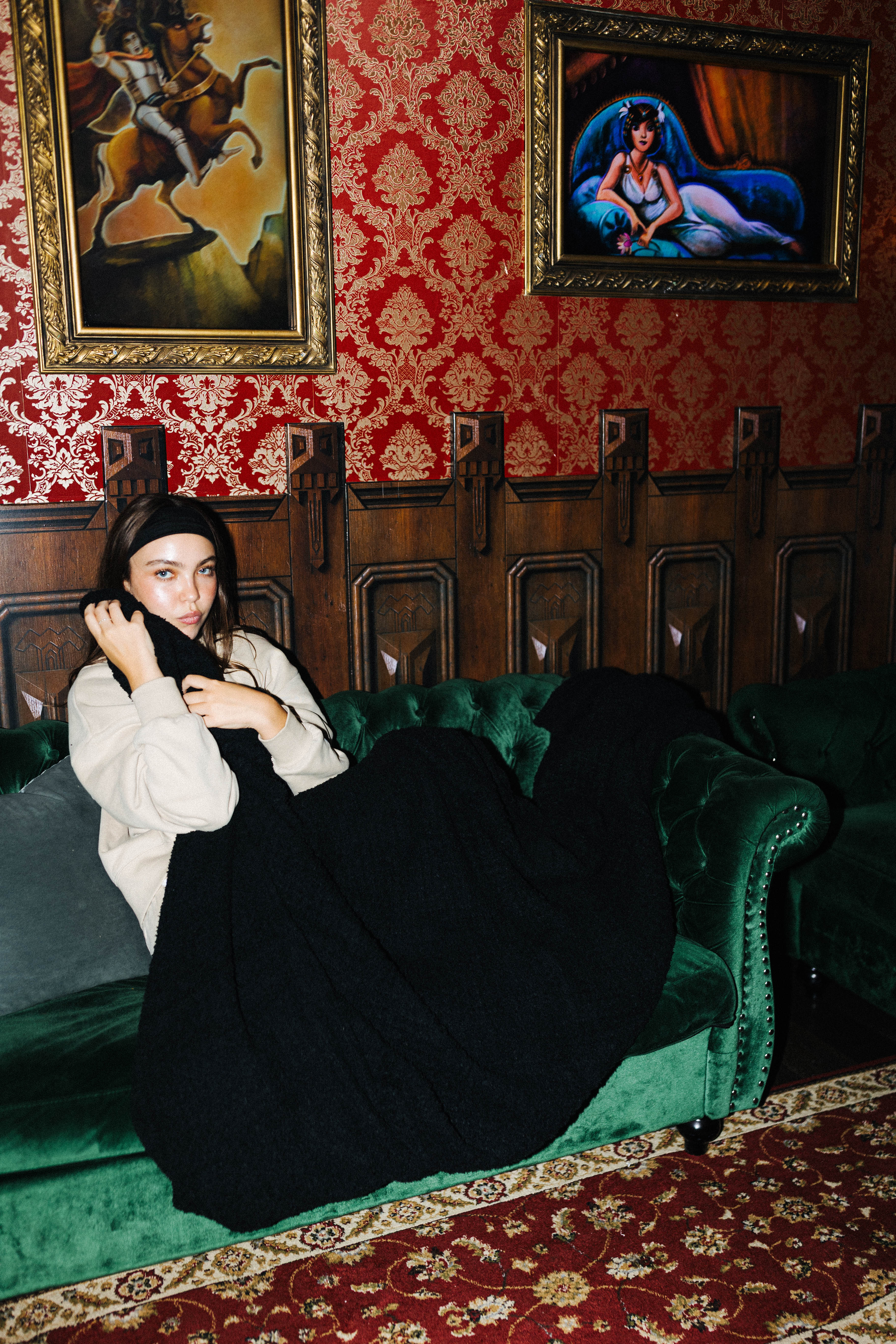 A woman reclines on a green velvet sofa in a vintage-style room with red patterned wallpaper, framed paintings, dark wood paneling, and a red patterned rug. She is wearing a cream-colored top and a long black skirt or dress.