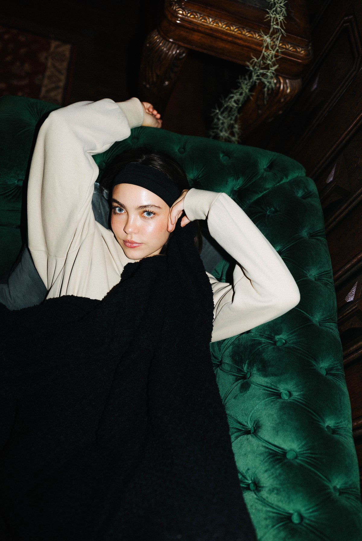 A model wearing a cream long-sleeve top, black headband, and black knit sweater reclines on a dark green tufted velvet sofa in a dimly lit room.