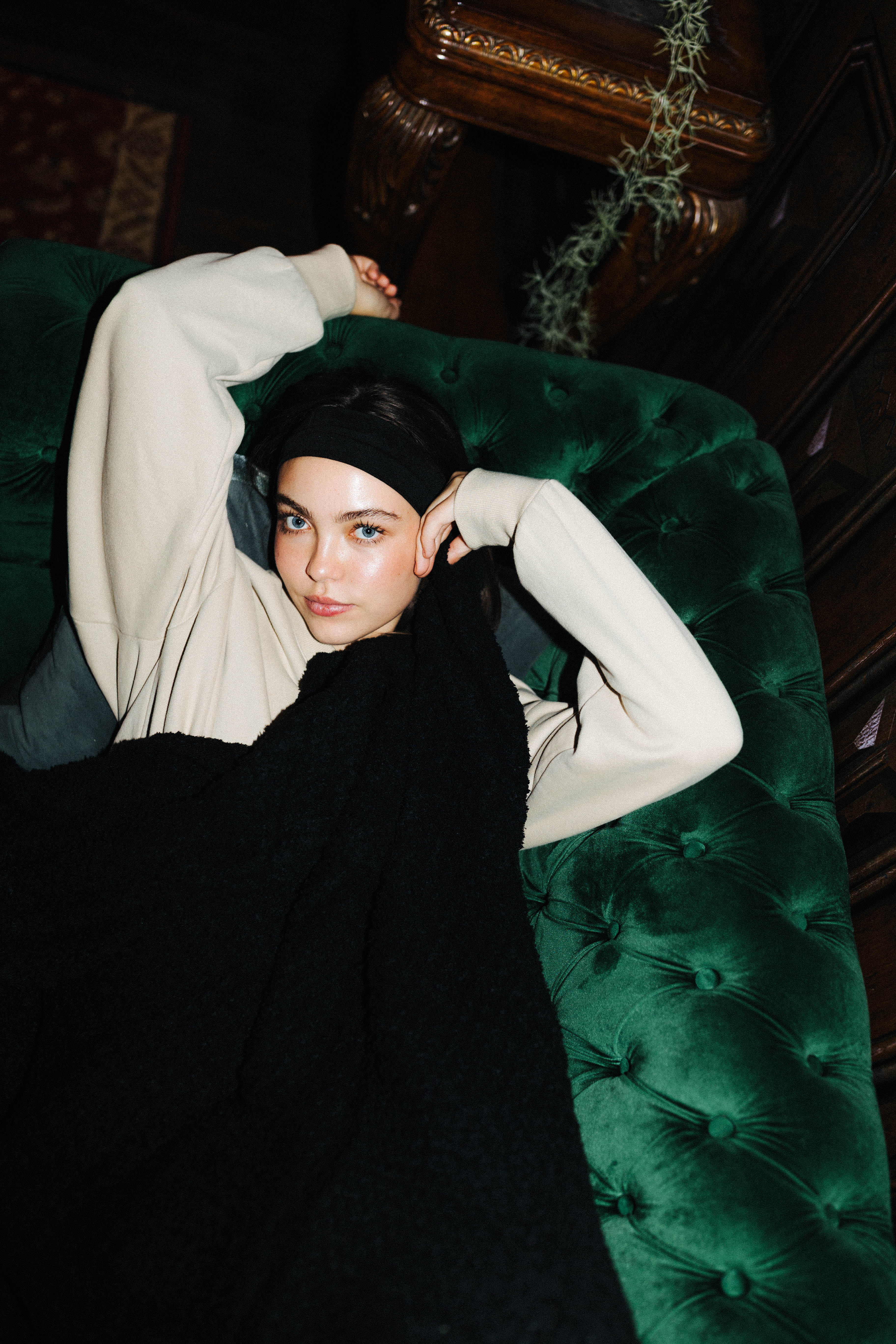 A model wearing a cream long-sleeve top, black headband, and black knit sweater reclines on a dark green tufted velvet sofa in a dimly lit room.
