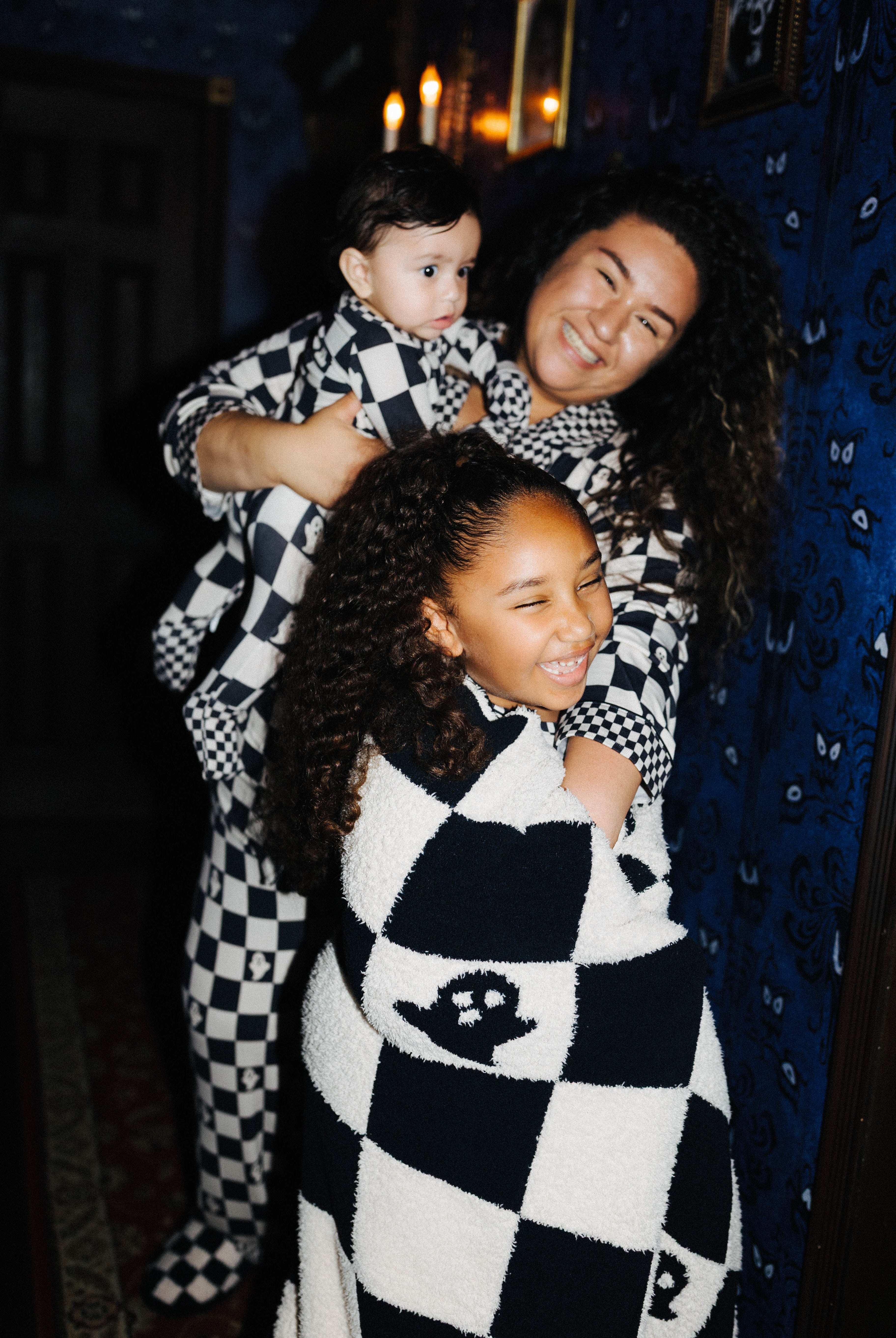 A woman and two children wearing matching sand-and-white checkered cozy pajamas or blankets in a dimly lit room with blue patterned wallpaper and warm candlelight.