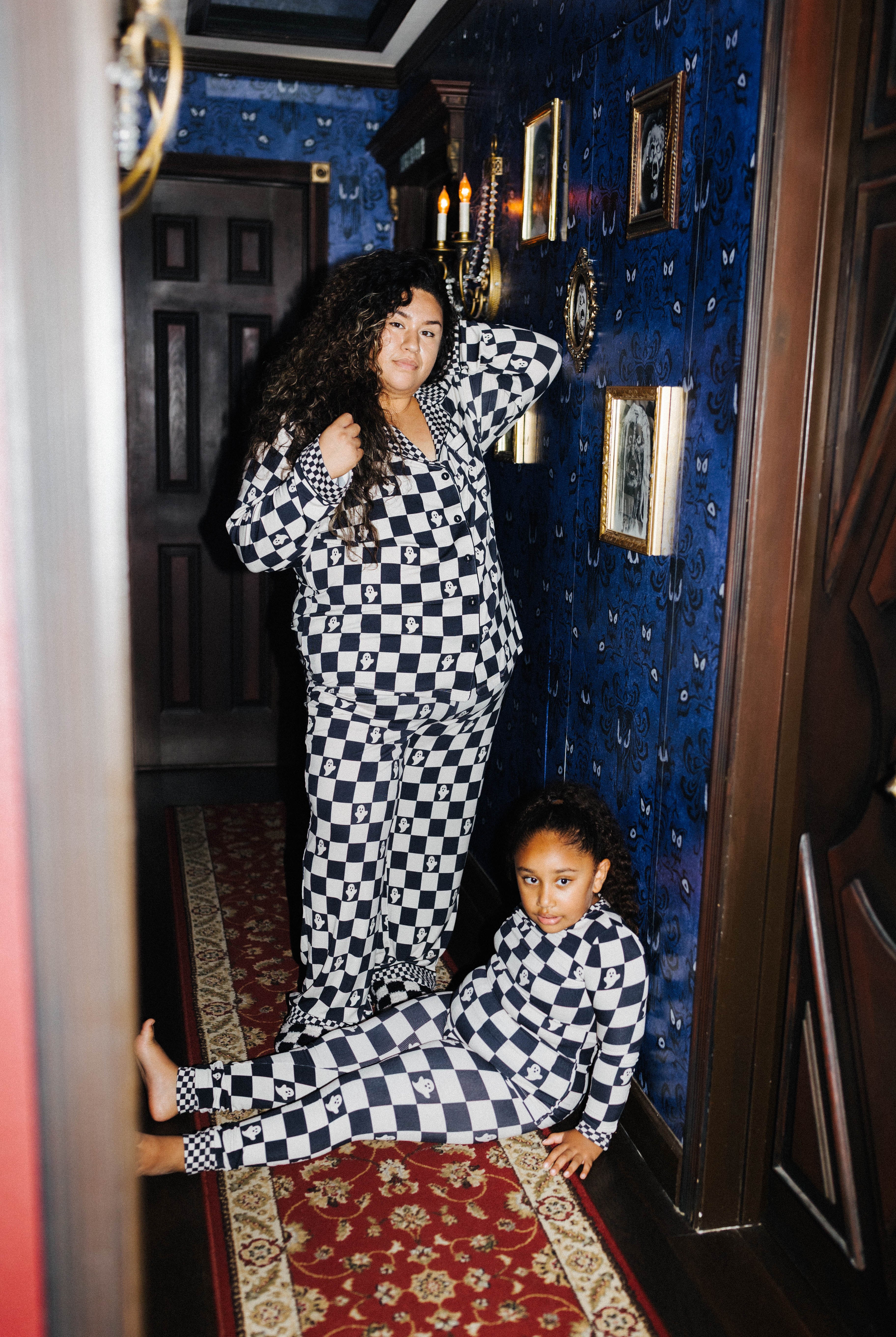 A woman and a child wearing matching black and white checkered pajamas pose in a dark, vintage-style room with blue patterned wallpaper and a red rug.