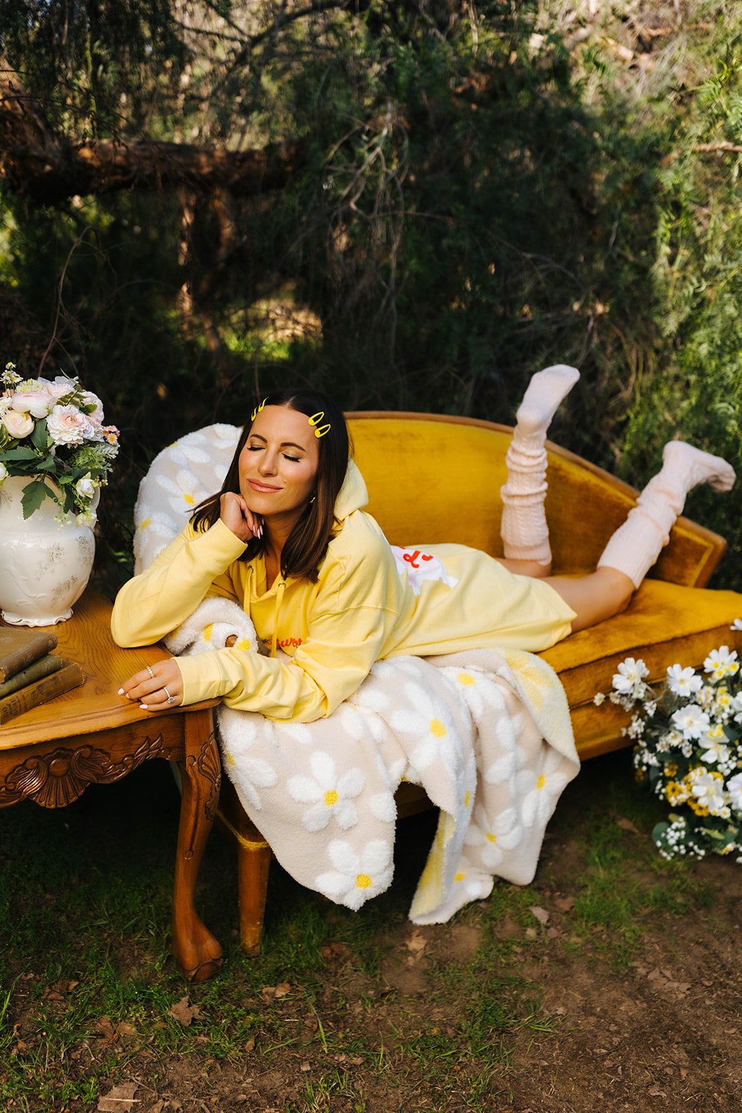 Woman in a yellow outfit sitting on a wooden bench outdoors with flowers and greenery around.