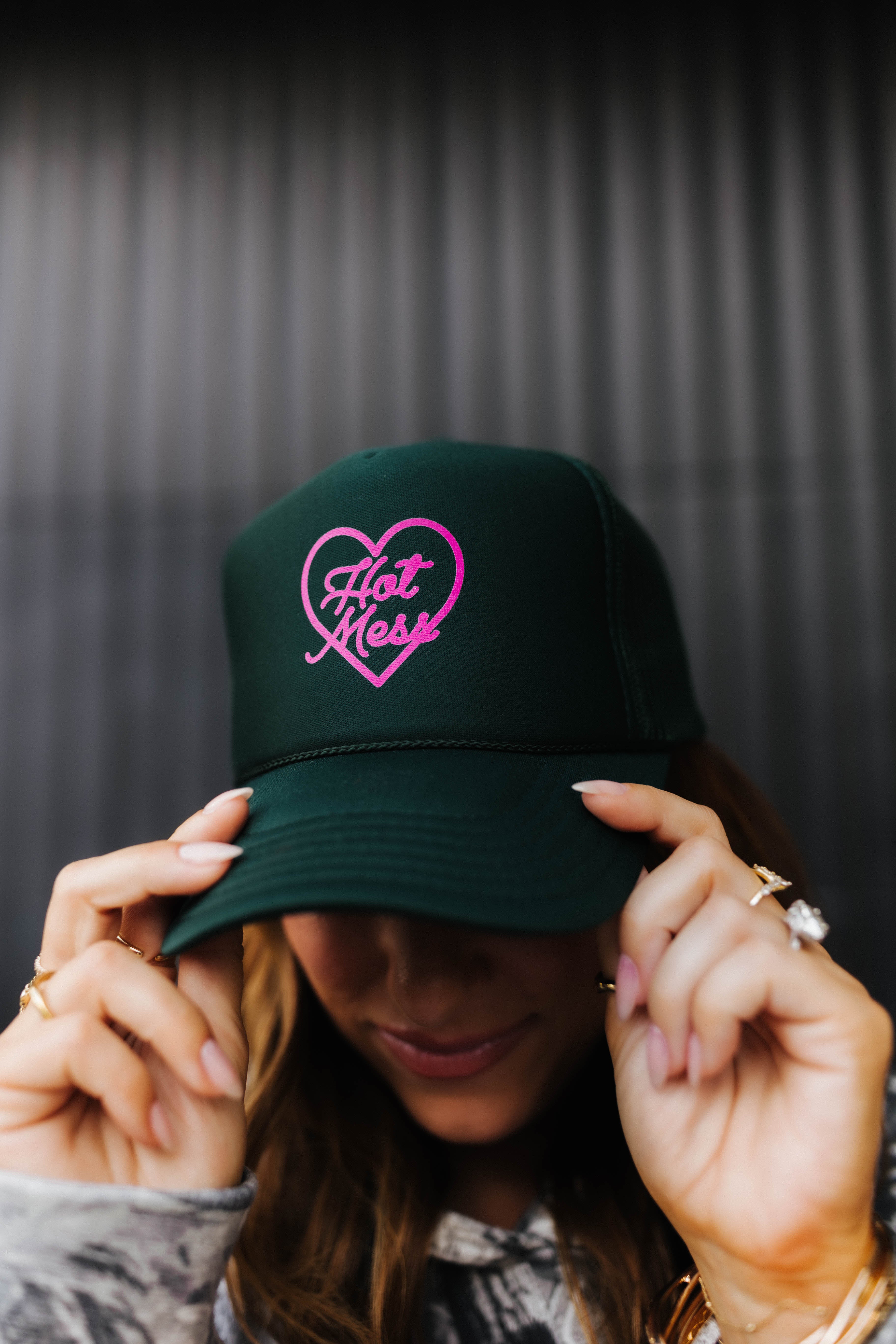 dark green hot mess at heart hat on model holding the bib of the hat with pink text and pink heart