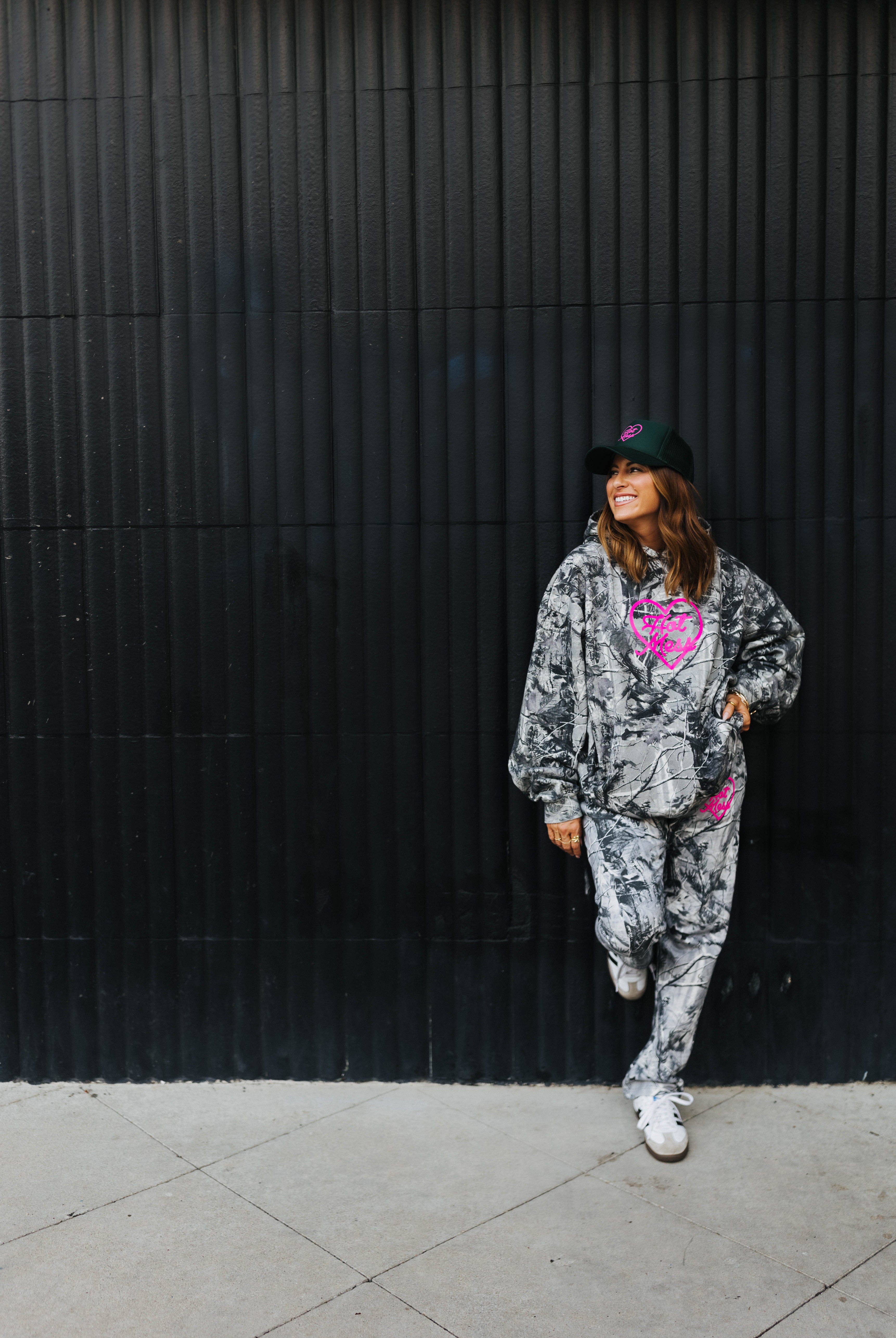 model leaning against black wall smiling wearing hot mess at heart camo set with hot mess at heart hat