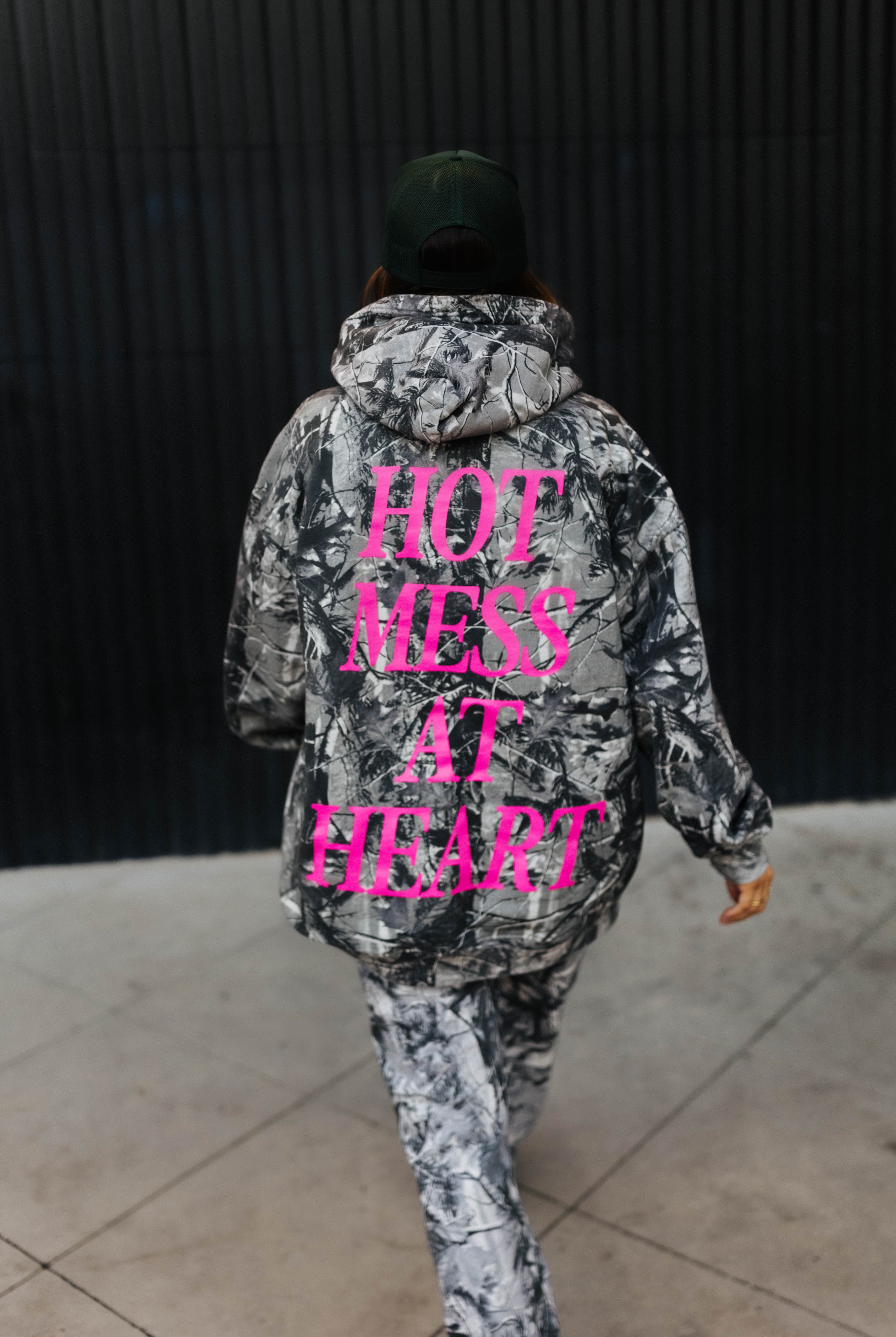 model walking while wearing hot mess at heart camo hoodie showing the back of the hoodie "hot mess at heart" in pink text