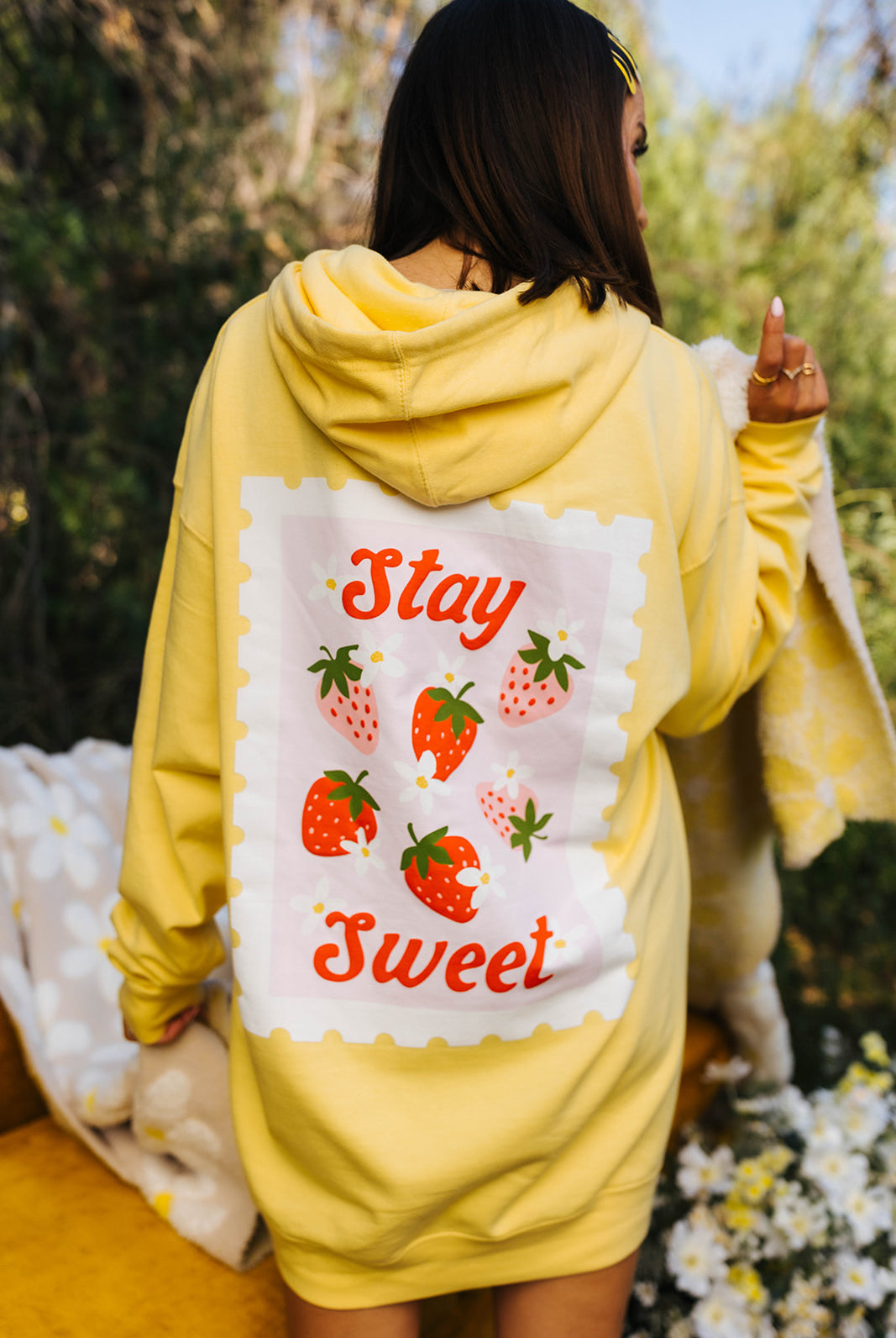 Person wearing a yellow hoodie with a 'Stay Sweet' design on the back, standing outdoors.