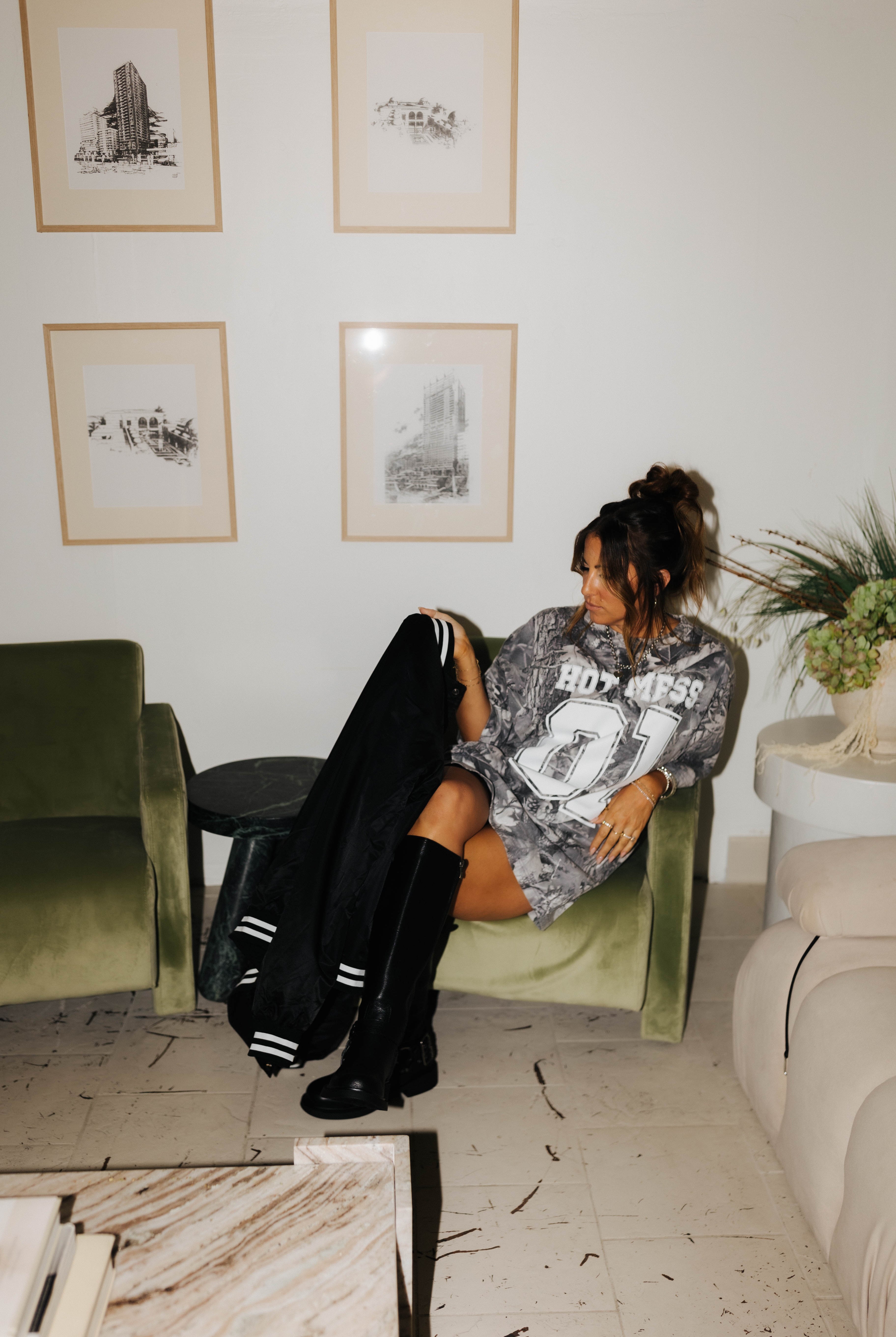 model sitting in chair wearing camo tee holding and looking at hot mess bomber jacket