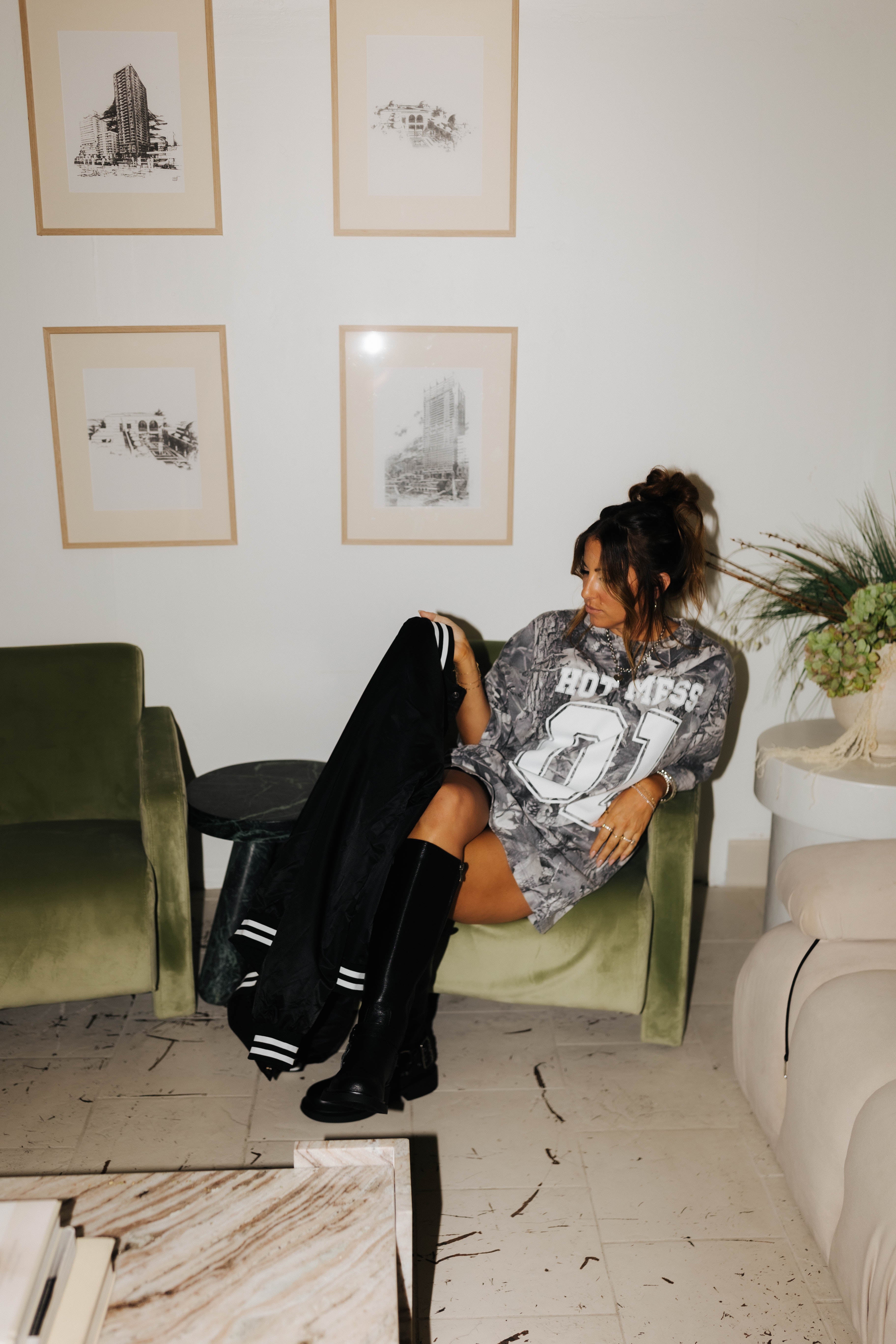 model sitting in chair wearing camo tee holding and looking at hot mess bomber jacket