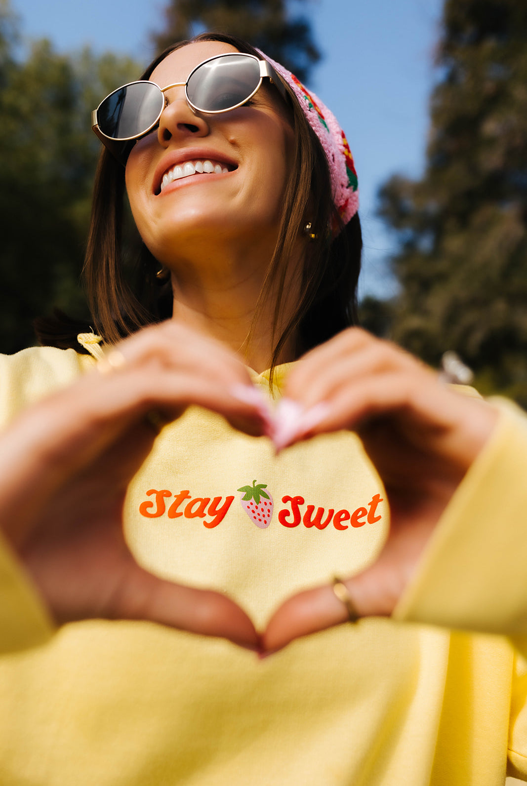 Woman in yellow sweatshirt and sunglasses making a heart shape with her hands, with 'Stay Sweet' text.