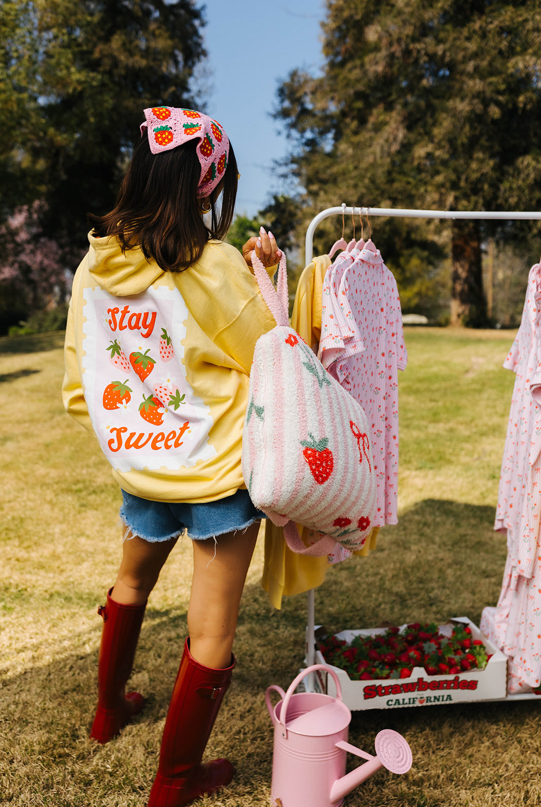 Person wearing a yellow hoodie with text, holding a pink bag with strawberry design, standing outdoors with clothes on a rack.