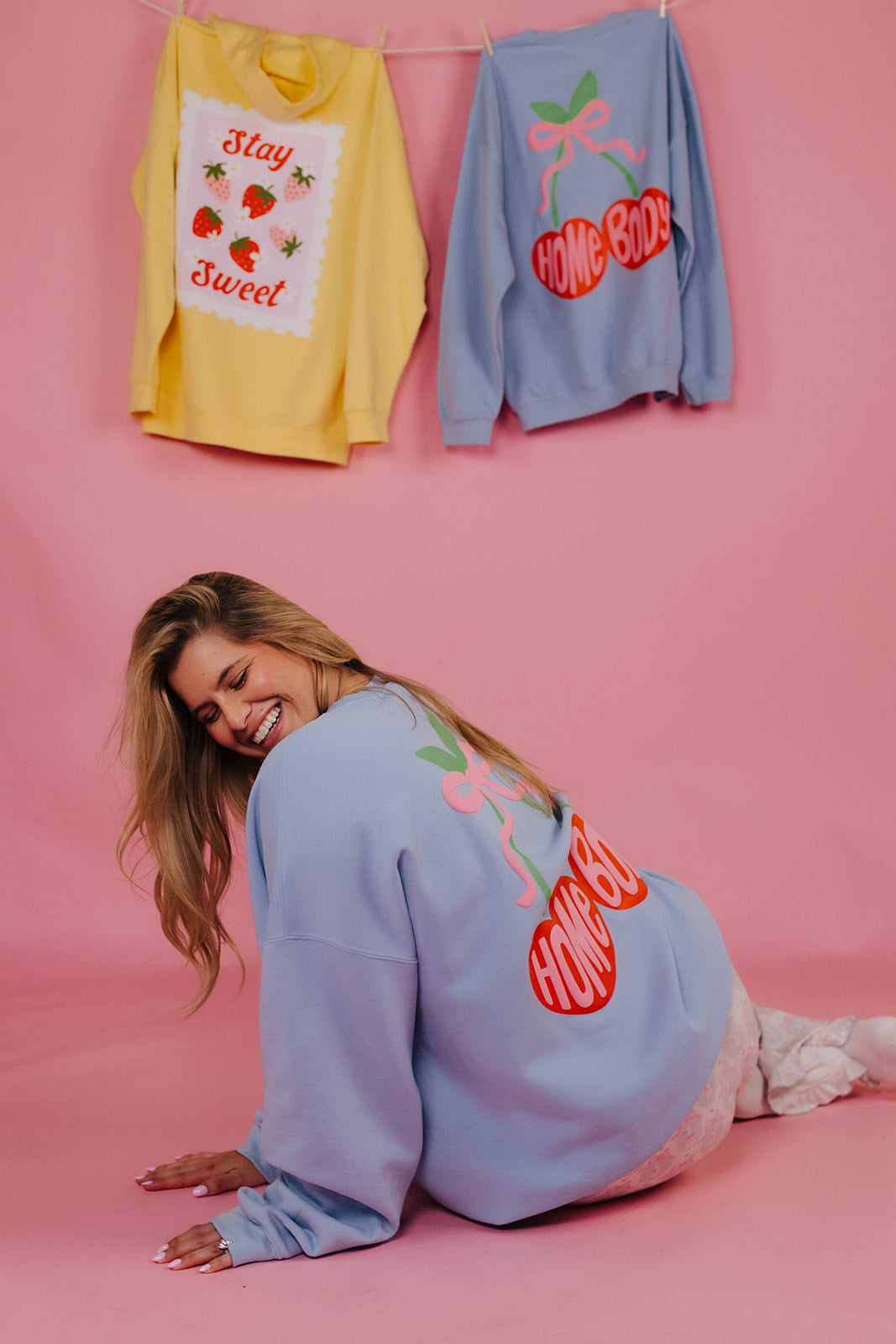 Woman wearing a light blue sweatshirt with red text and graphics, sitting on a pink background with two sweaters hanging above her.
