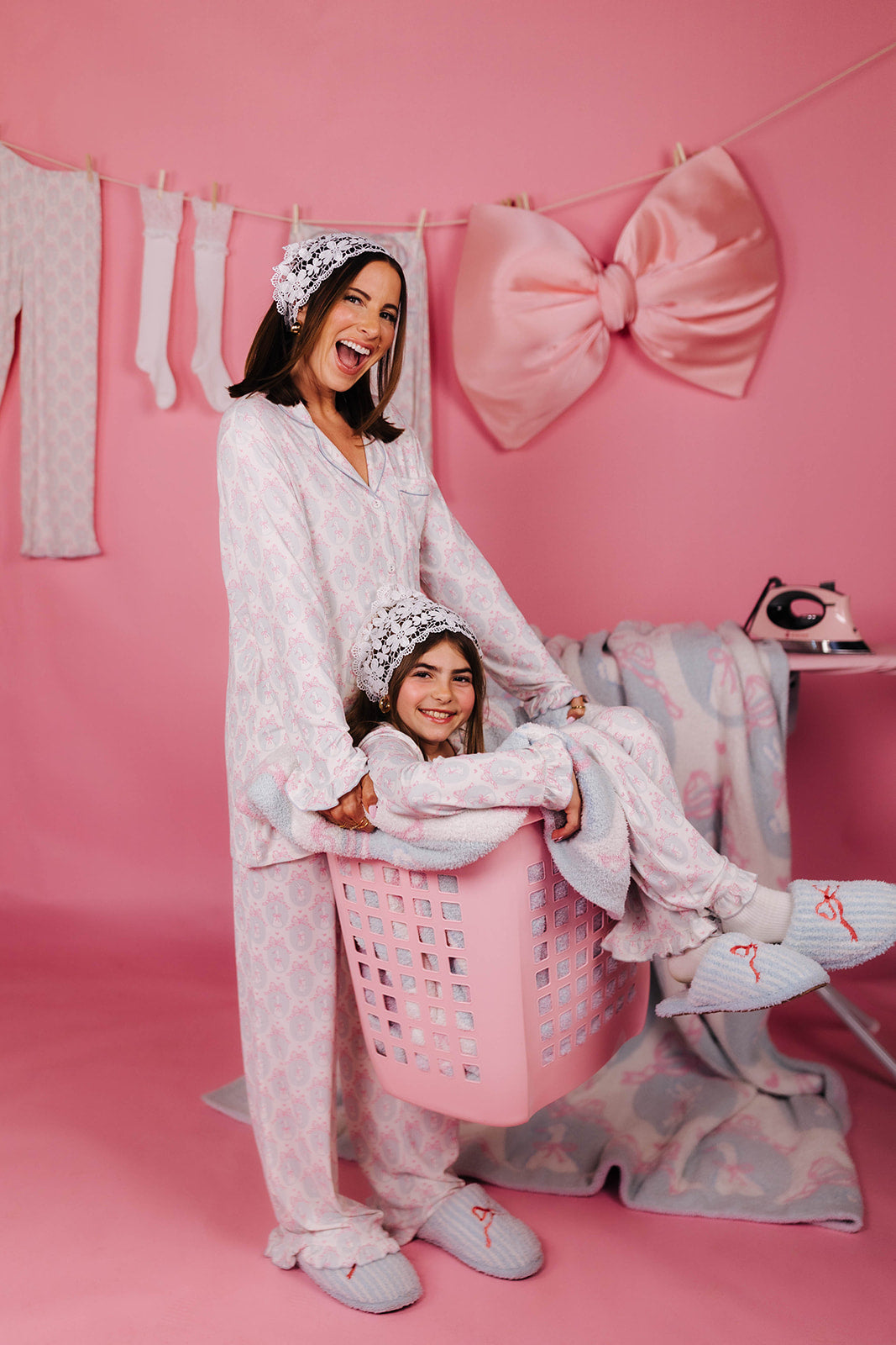 Woman and child in matching pajamas with a pink bow and laundry basket on a pink background
