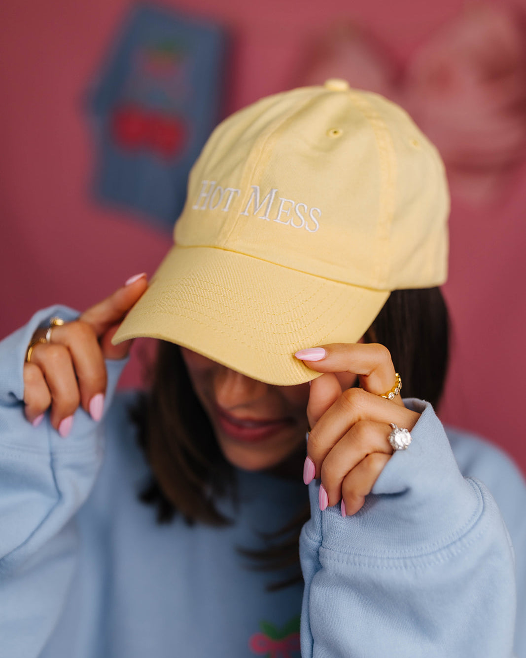 Person wearing a yellow cap with text, holding it over their face against a pink background hat says hot mess