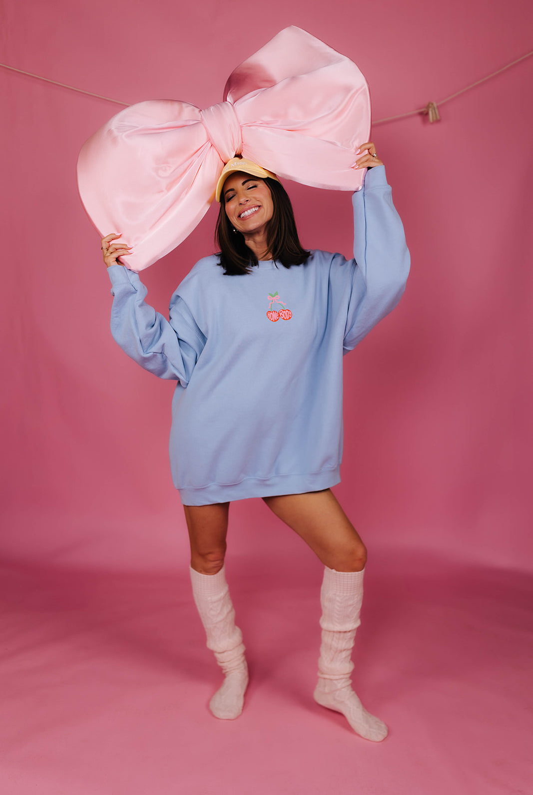 Person wearing a light blue oversized sweatshirt and white knee-high socks, holding a large pink bow against a pink background.