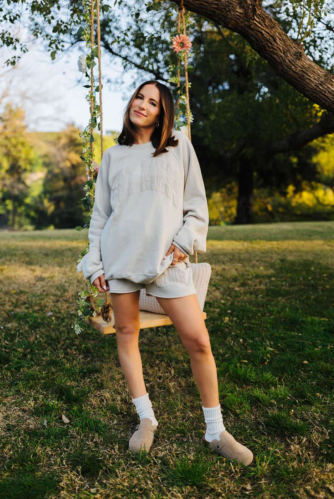 Woman in a light tan sweater and shorts sitting on a swing in a park.