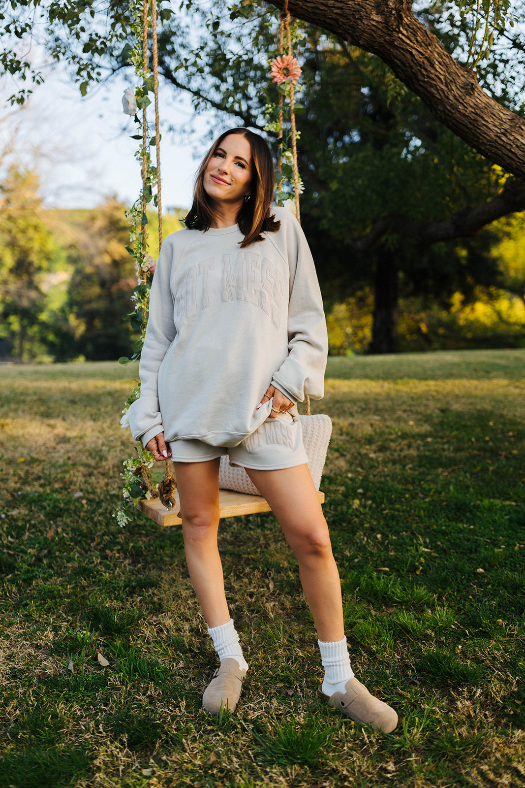 Woman in a light tan sweater and shorts sitting on a swing in a park.