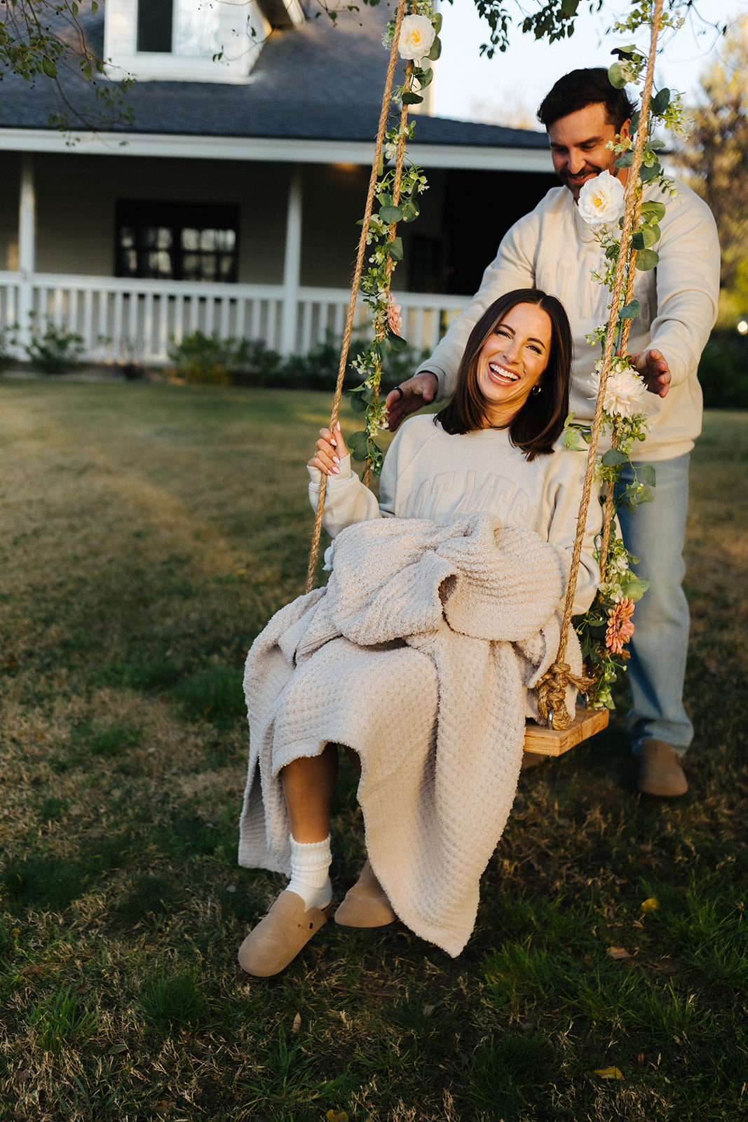 Woman sitting on a swing held by a man in a garden setting with a house in the background. holding the quillow unfolded