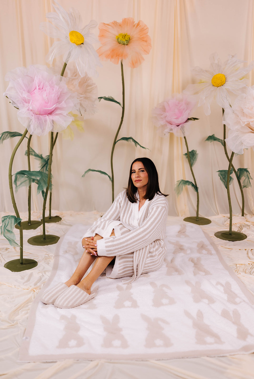 Woman sitting on a white bunny blanket surrounded by large decorative flowers in a softly lit room.
