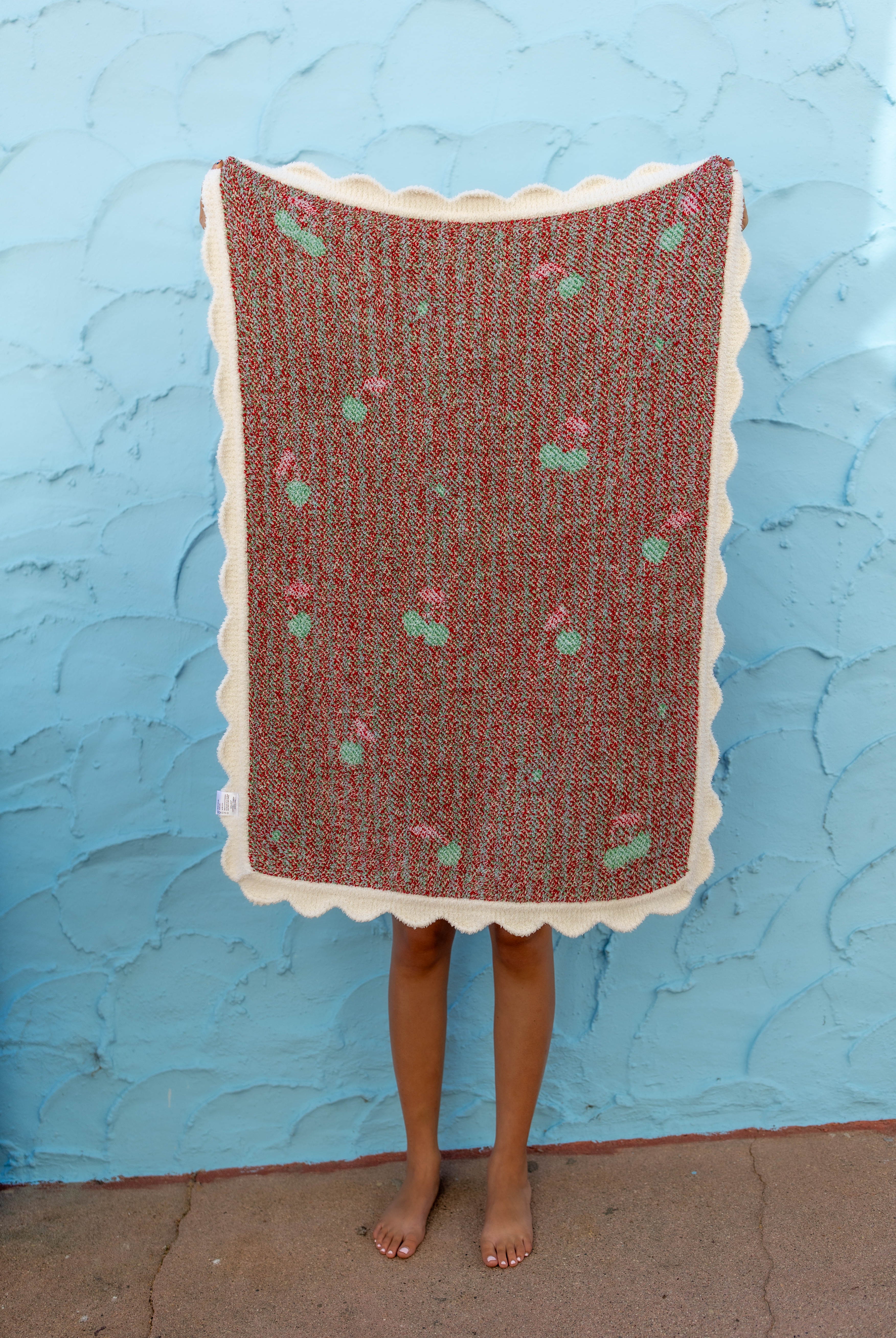 A person standing barefoot in front of a textured light blue wall, holding up a rectangular woven blanket with a scalloped cream border.