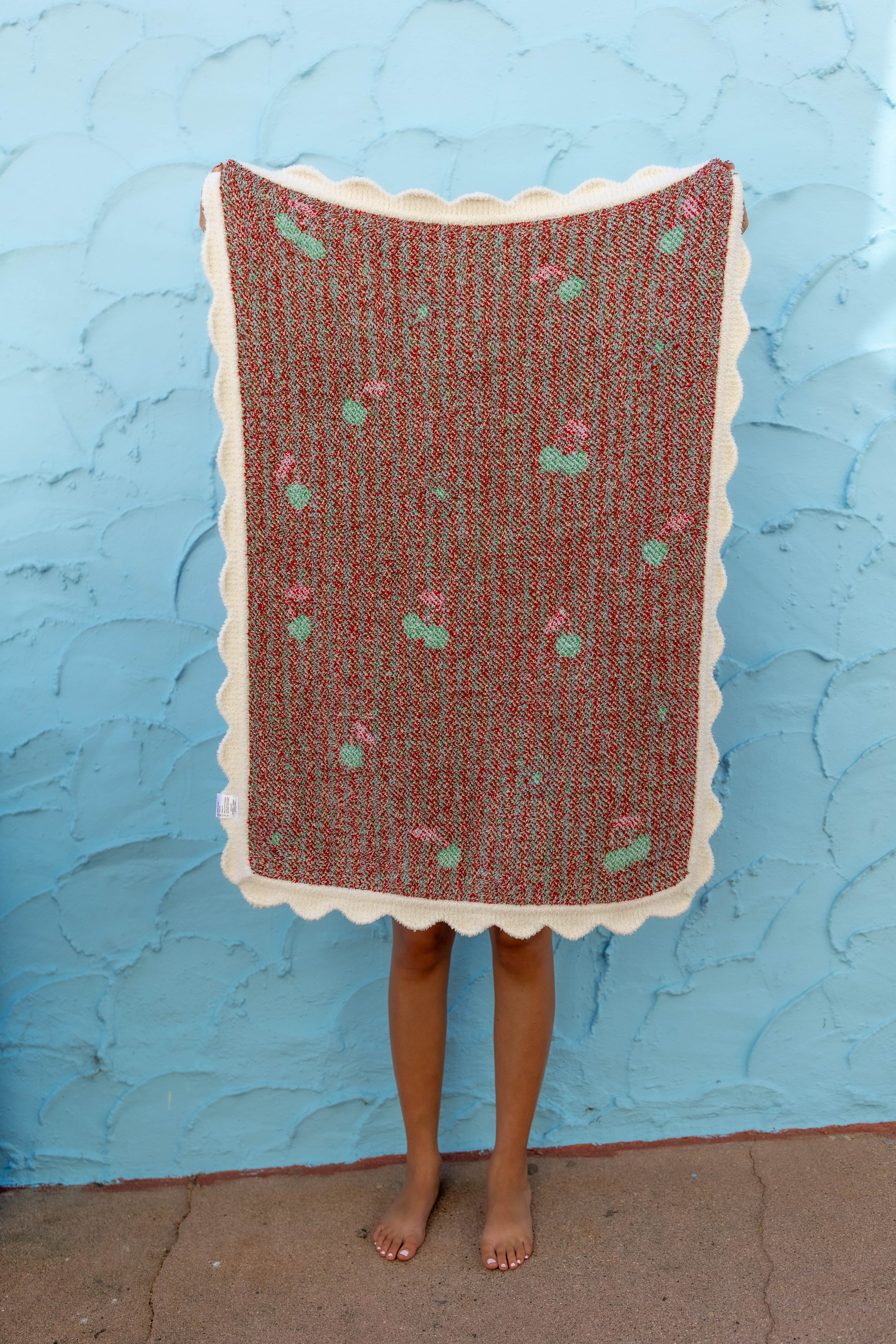 A person standing barefoot in front of a textured light blue wall, holding up a rectangular woven blanket with a scalloped cream border.