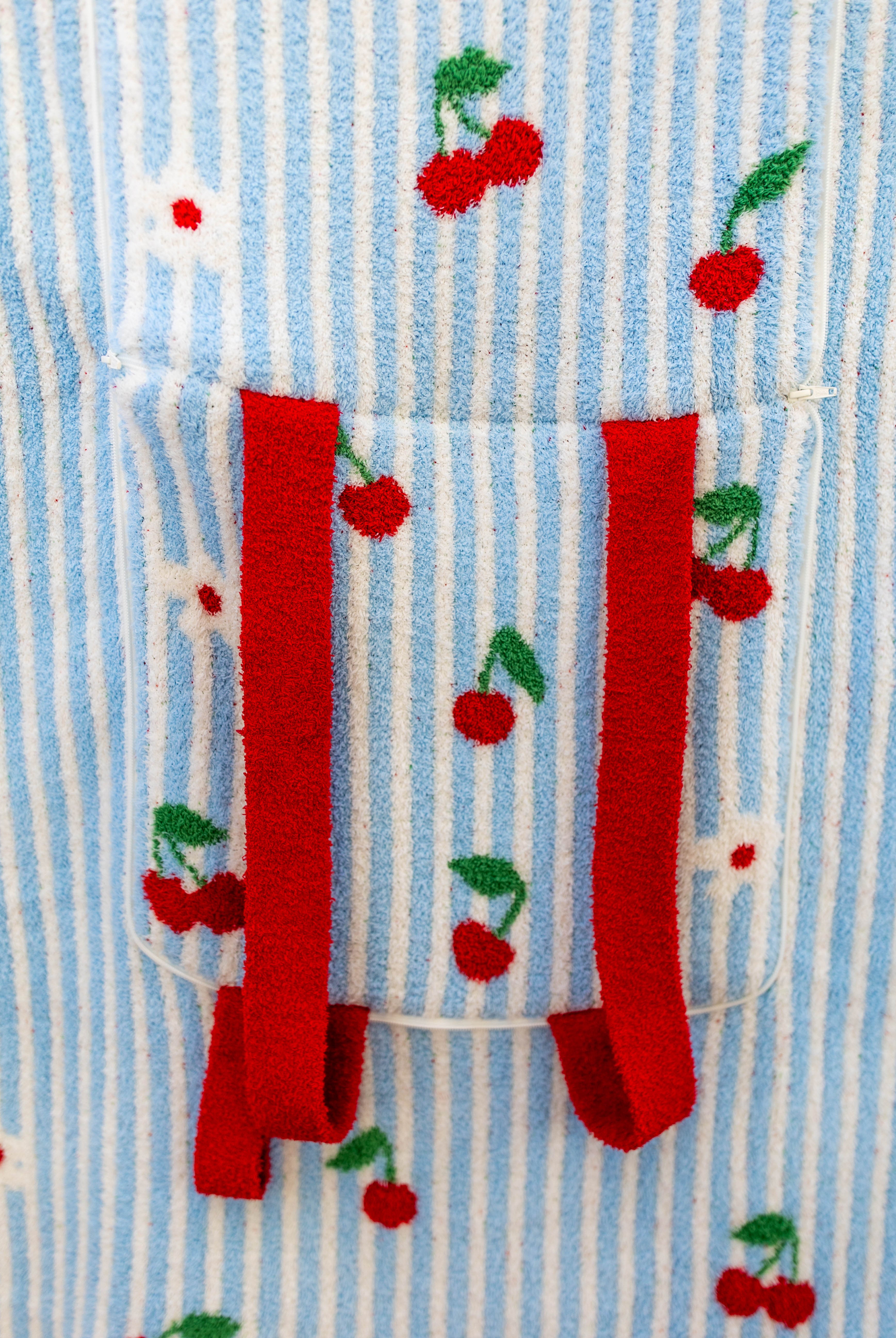 Close-up of a light blue and white striped fabric with embroidered red cherries and green leaves, featuring a front pocket with red straps.