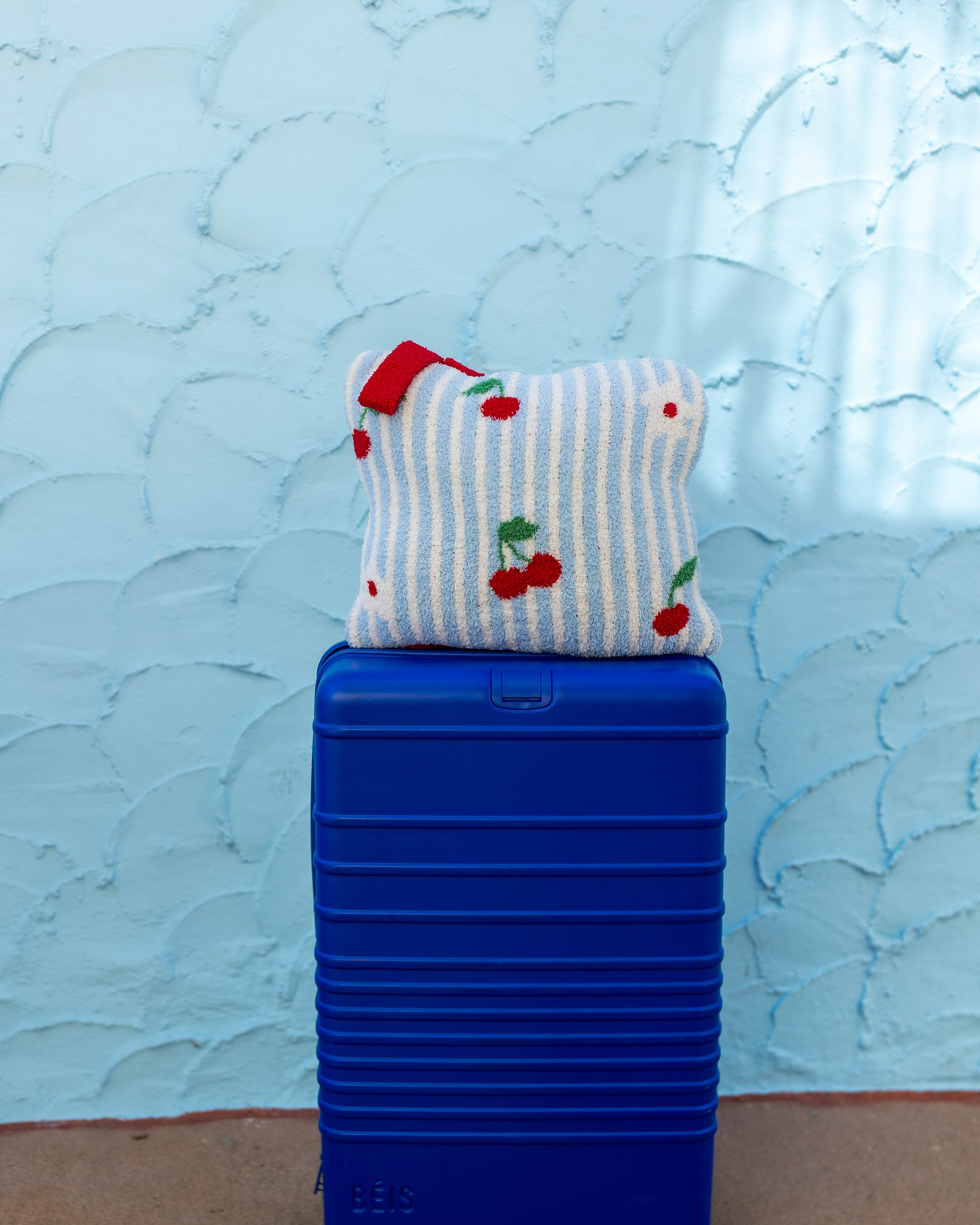 A bright blue hard-shell suitcase standing upright on a tan floor against a light blue textured wall, with a striped and cherry-patterned quillow resting on top.