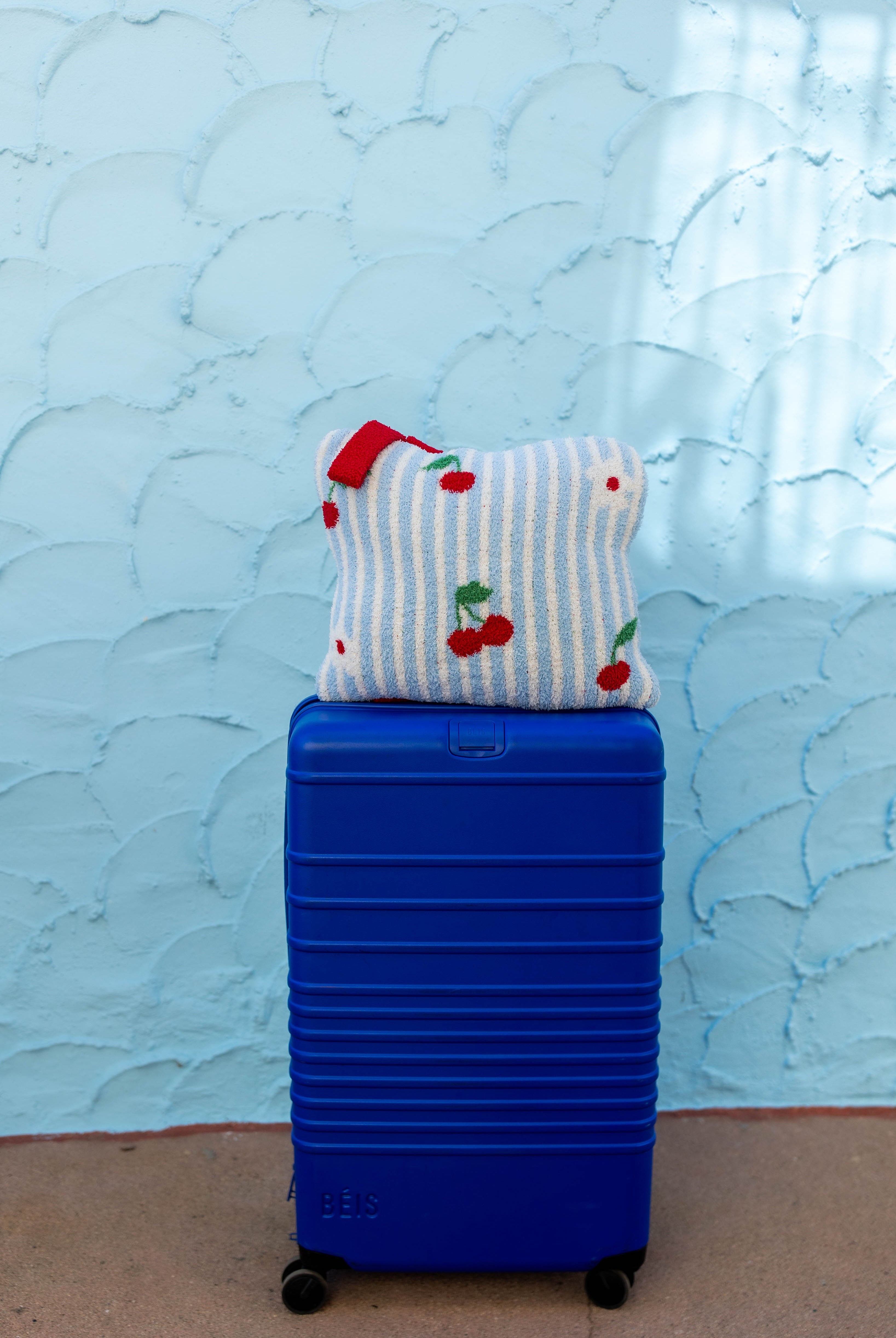 A bright blue hard-shell suitcase standing upright on a tan floor against a light blue textured wall, with a striped and cherry-patterned quillow resting on top.
