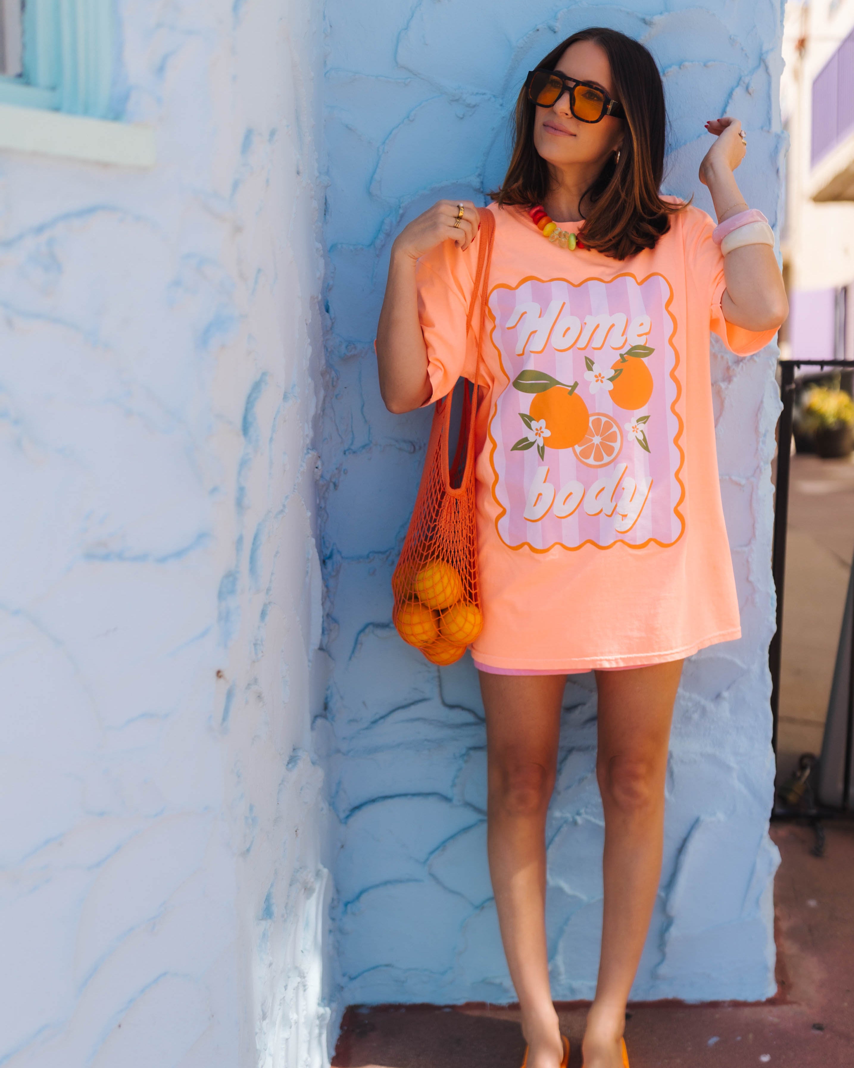 A woman stands outdoors against a light blue textured wall wearing an oversized orange graphic T-shirt with orange fruit illustrations and the words 'Home body,' along with orange flip-flops and a mesh tote bag filled with oranges.